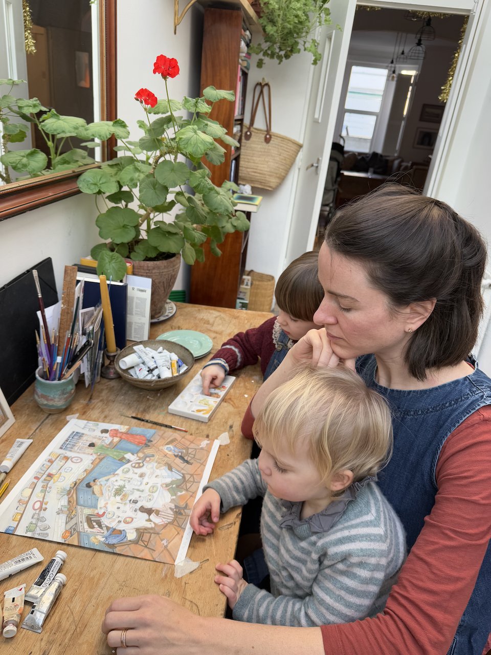 Anna Neima and two young children at a table, looking at a children's puzzle or book. The table has art supplies, tubes of paint, and a potted plant with red flowers nearby. The scene is inside a cozy home with a mirror, bookshelf.
