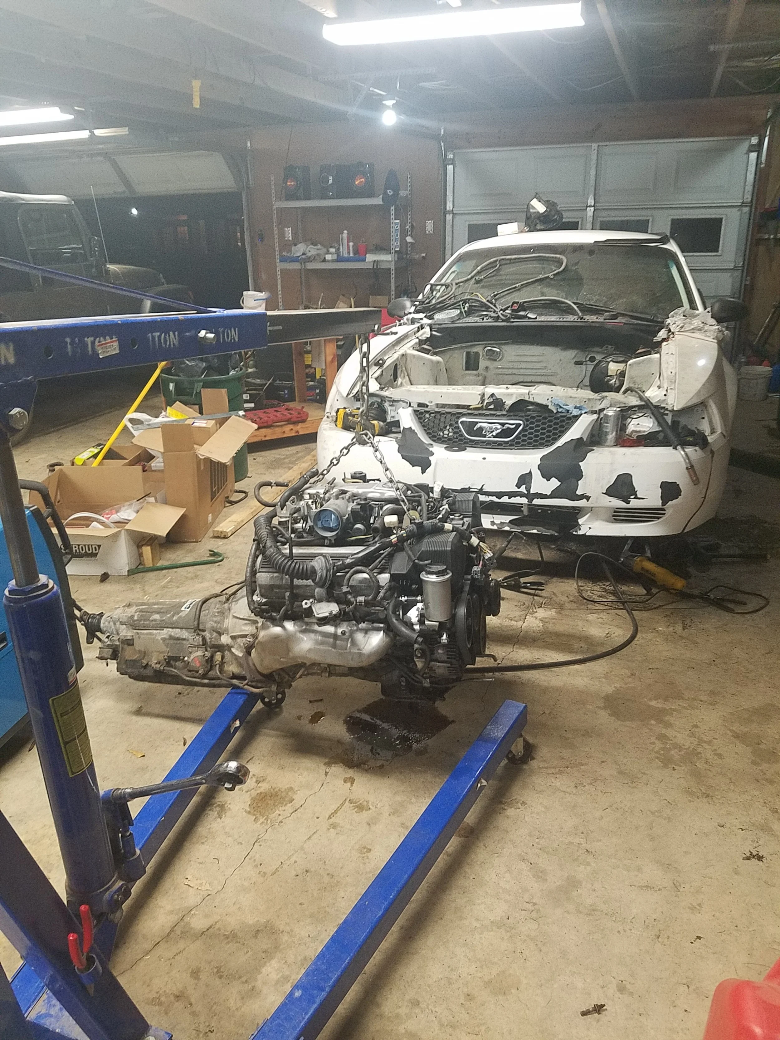 In a garage, a white 24 hours of lemons racecar, the Lustang, with front damage is being repaired. The hood is open, and the engine is removed. An engine is suspended in the air in front of the car, supported by a blue engine hoist.