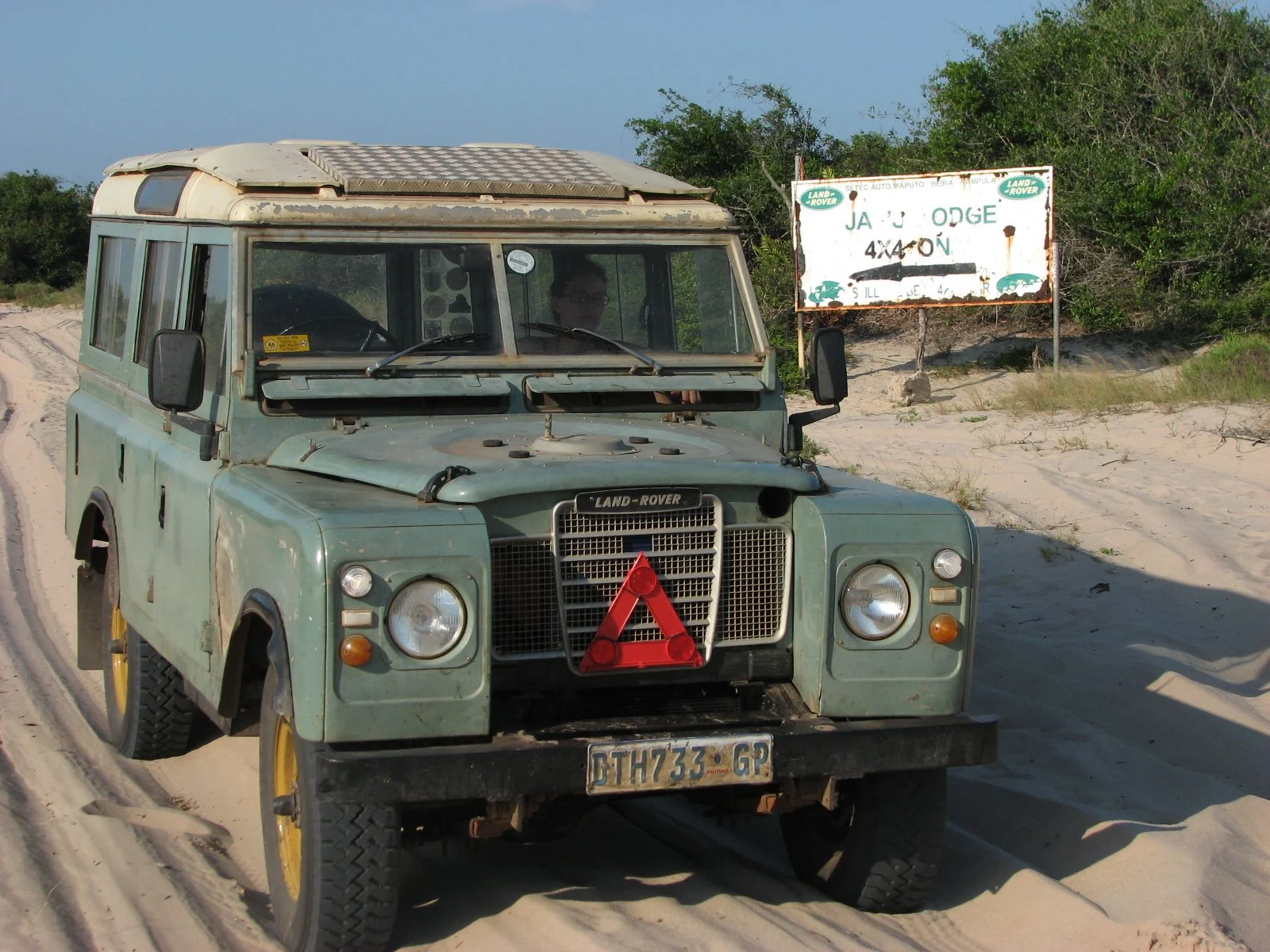 An old, weathered green Land Rover Series 3 parked on sandy terrain in Mozambique with a rusty signboard in the background that reads "Jano Lodge 4x4 On."
