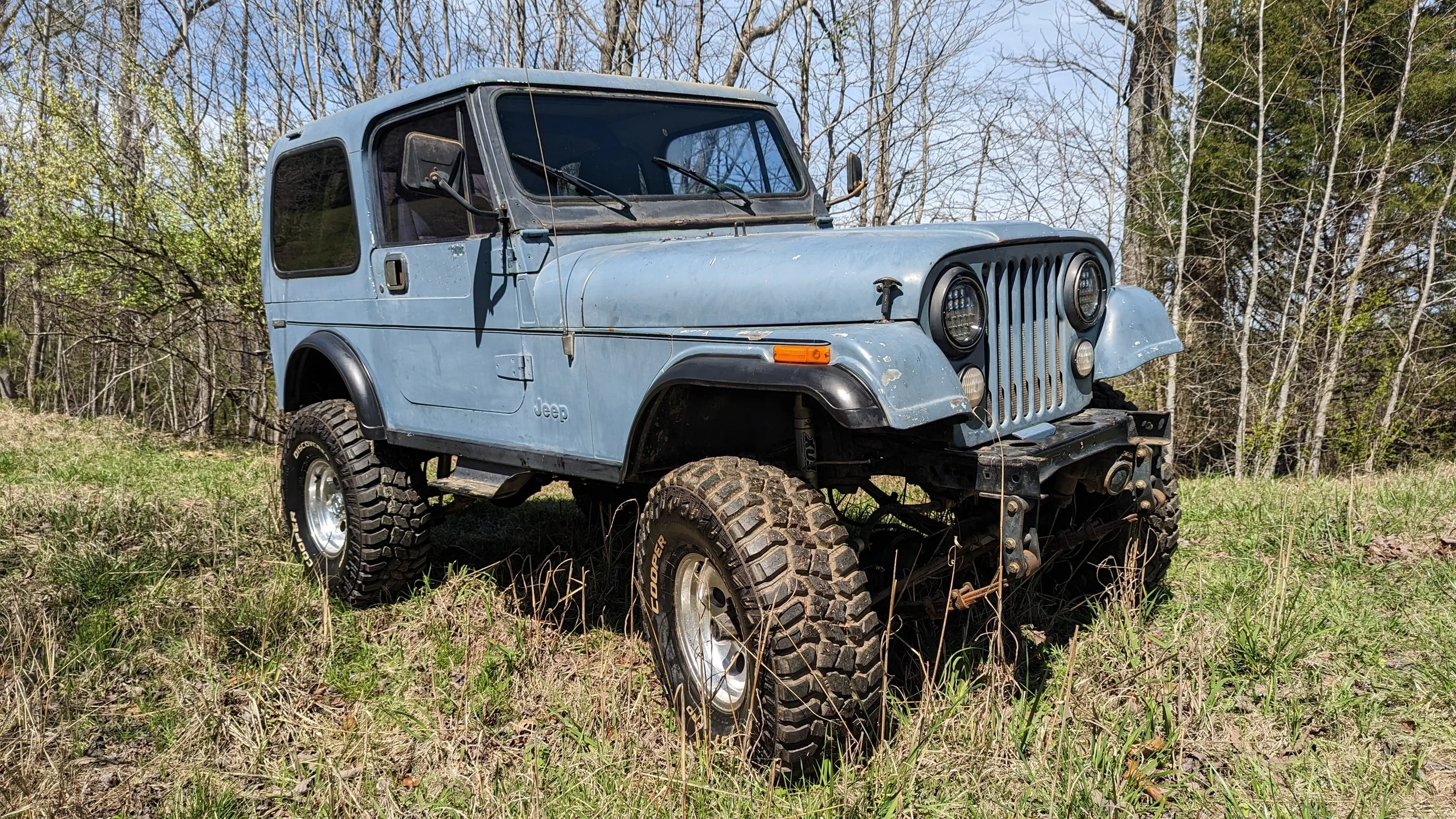 A vintage blue CJ7 Jeep with large 35" off-road tires a straight 6 4.0, 4"lift, parked in a grassy field with trees in the background.
