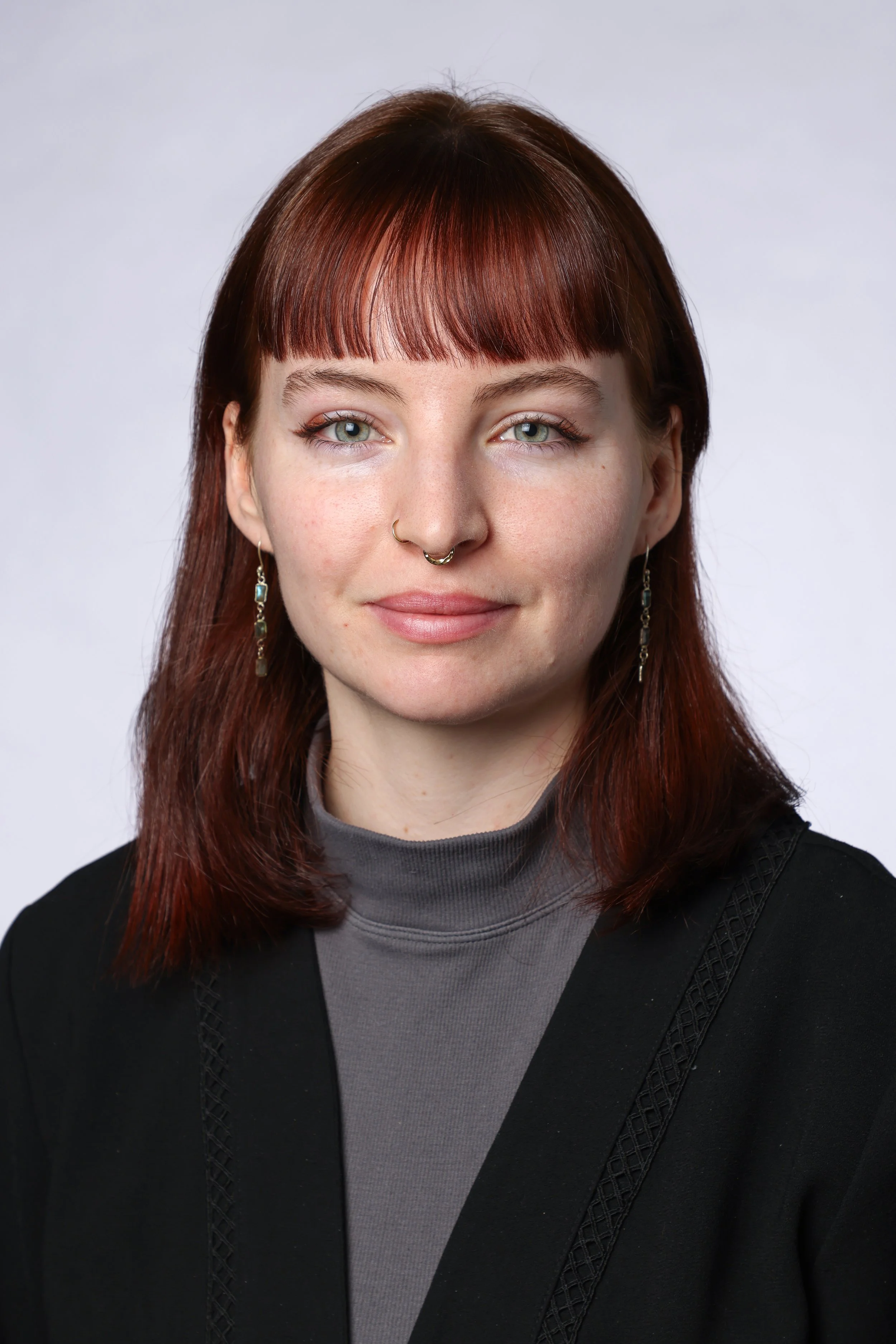 Portrait d'une femme aux cheveux roux avec des yeux clairs, portant un haut gris et une veste noire,avec des boucles d'oreilles et un piercing au nez, devant un fond blanc.