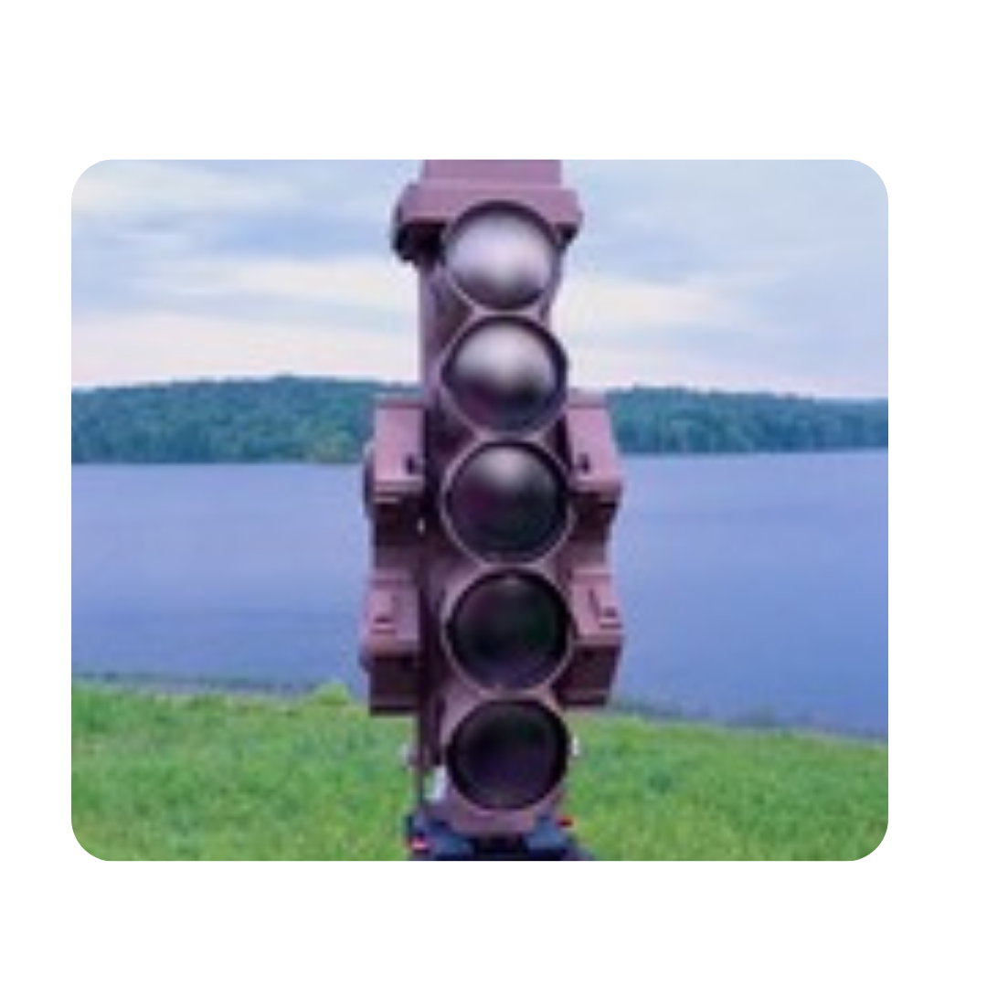 A giant traffic light sculpture with five circular sections, situated outdoors near a body of water with a grassy area in the foreground and trees in the background.