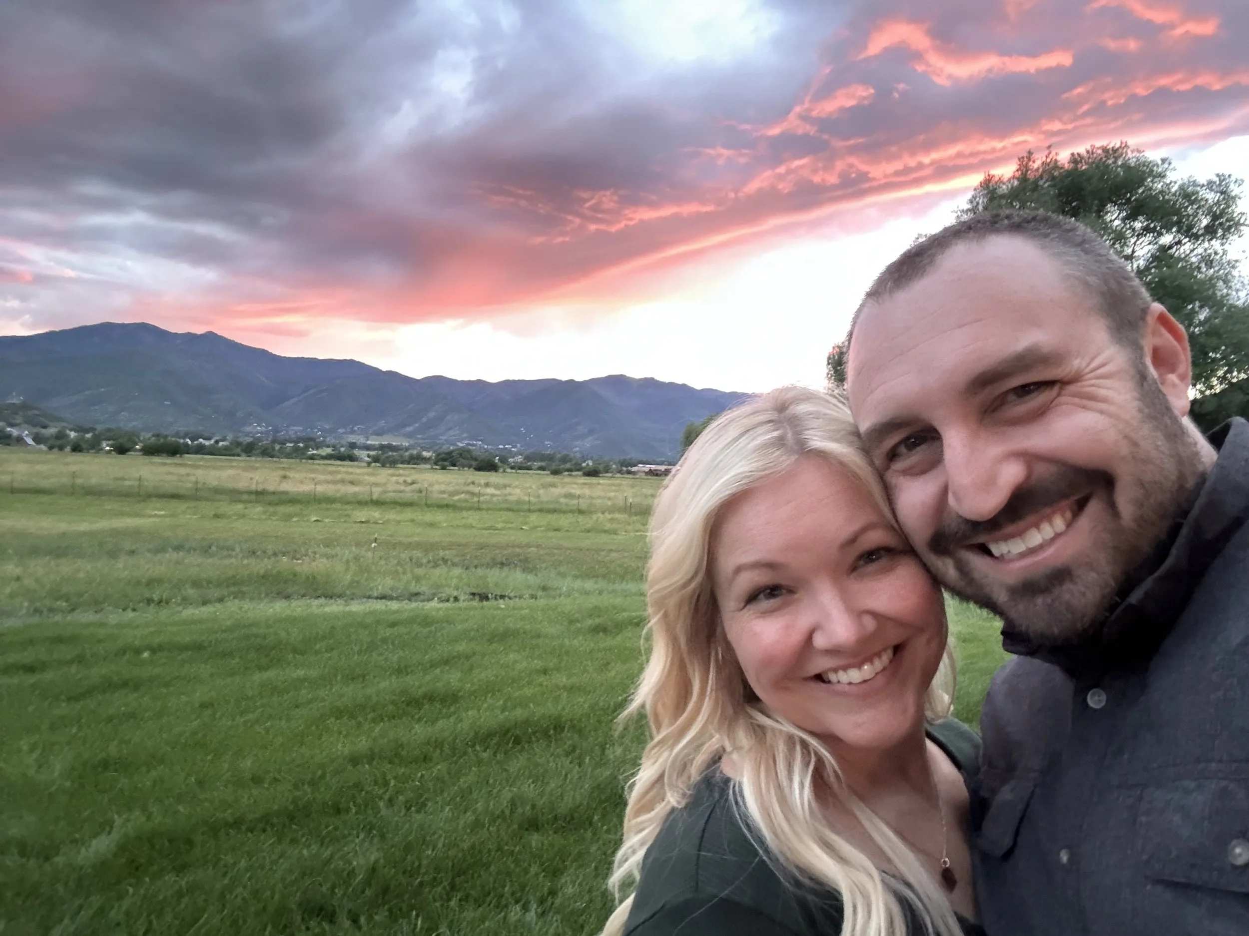 A smiling couple taking a selfie outdoors at sunset with mountains and a colorful sky in the background.