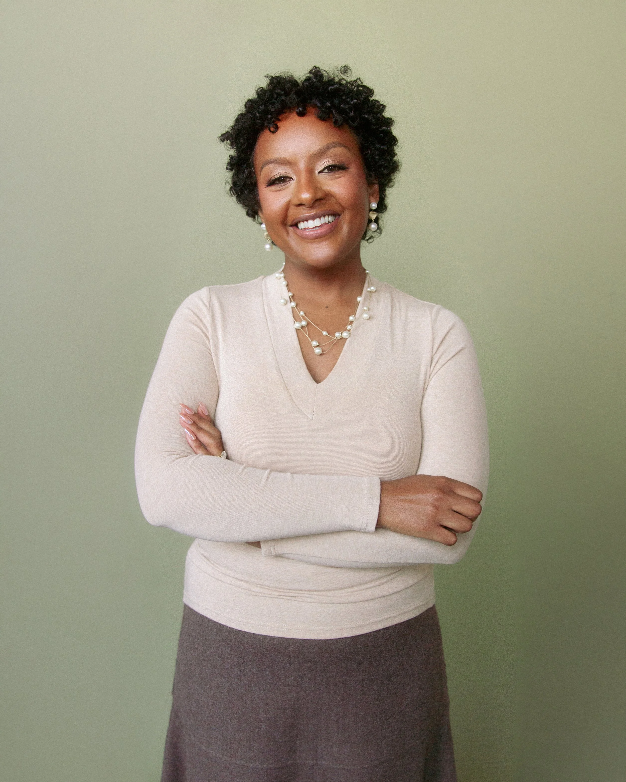 A smiling woman with short curly black hair, dressed in a beige long-sleeve top and a brown skirt, standing with arms crossed in front of a green background.