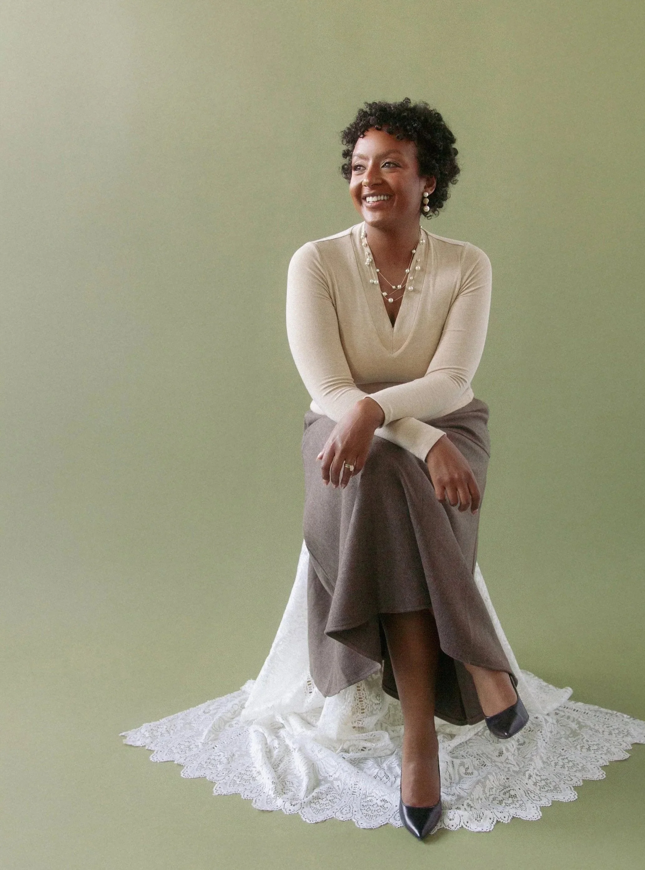 A woman with curly dark hair, smiling, wearing a beige top, gray skirt, black heels, and pearl jewelry, sitting on a white lace-covered stool against a light green background.