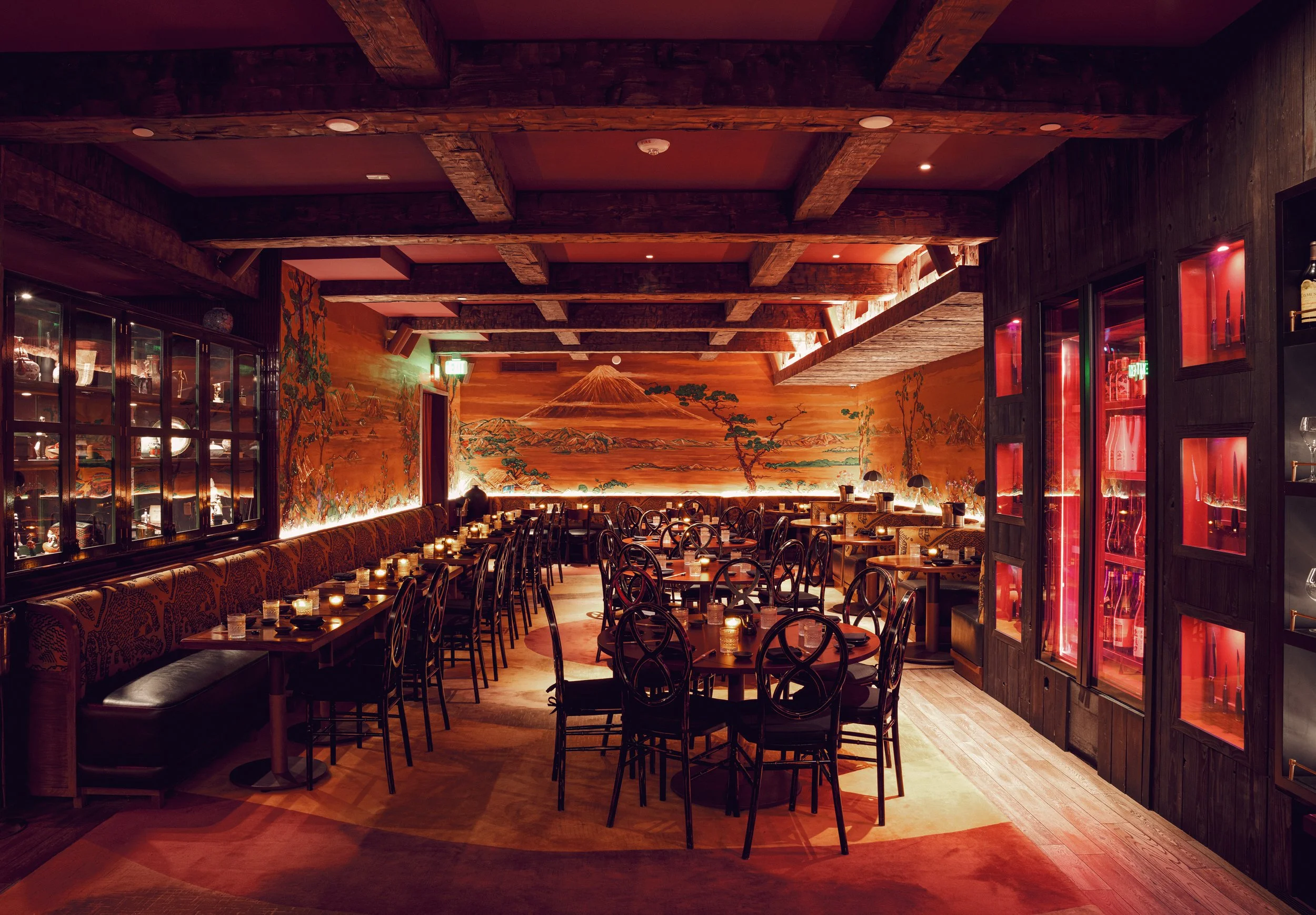Interior of a dimly lit restaurant with wooden furniture, a mural of a volcano landscape on the back wall, and shelves with wine bottles. There are tables set with candles, and the overall ambiance is warm and cozy.