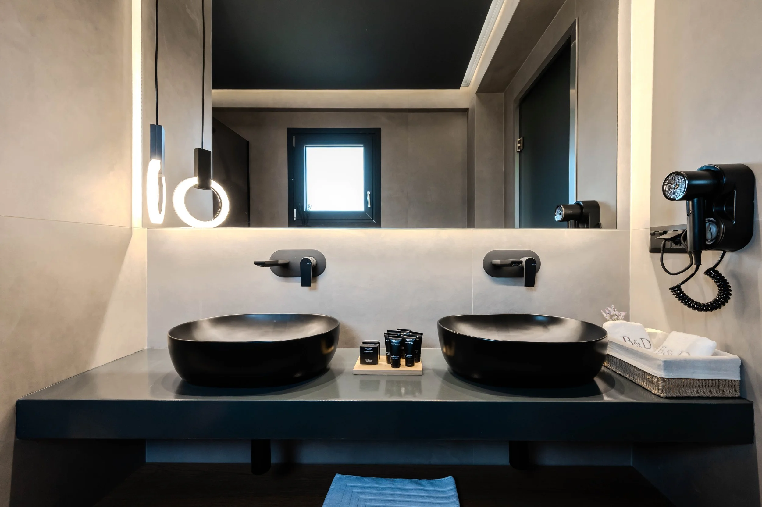 Modern bathroom with two black vessel sinks on a gray countertop, wall-mounted black faucets, a large mirror, a window, and wall-mounted hair dryer, with decorative toiletries and towels.