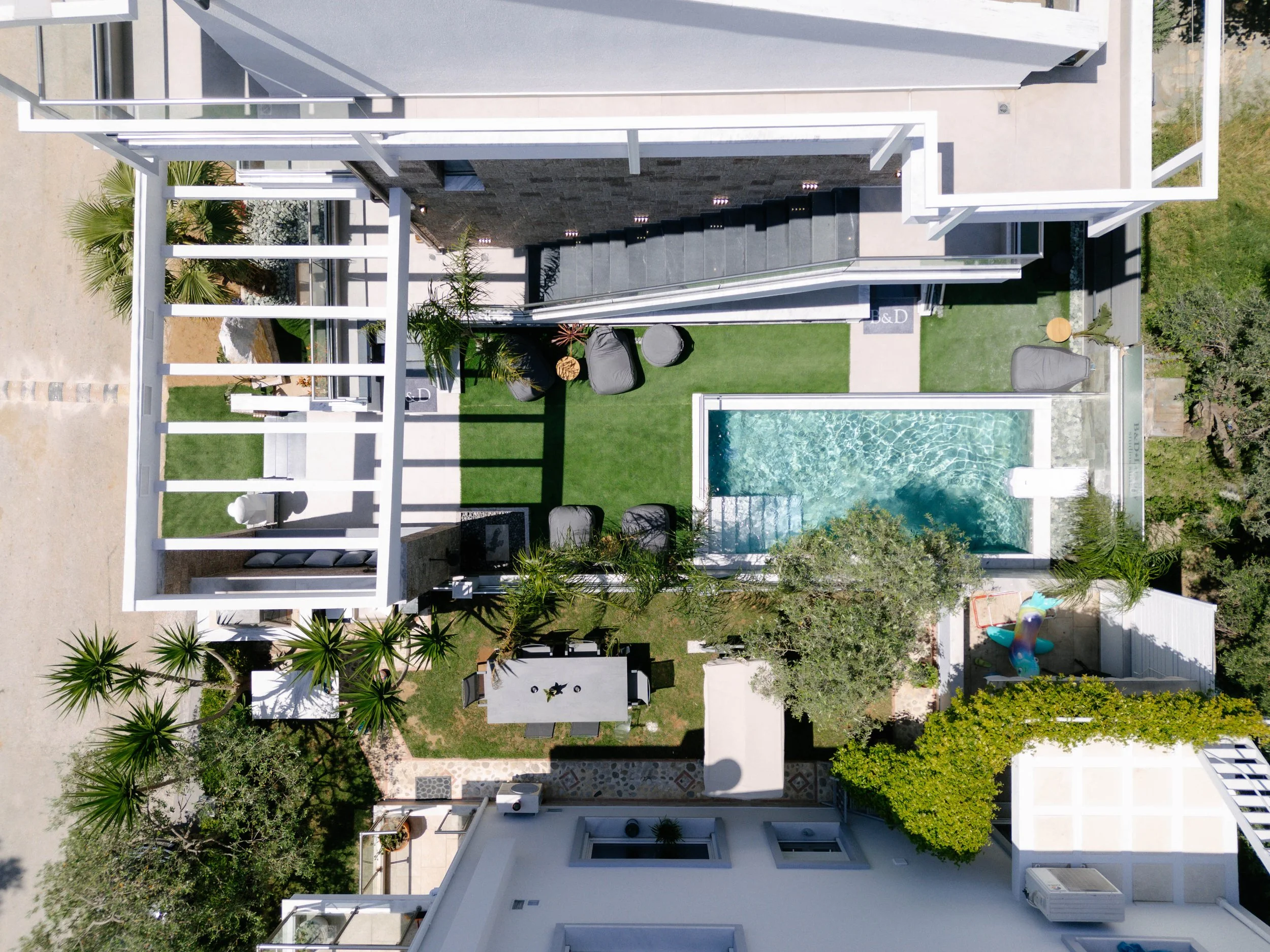 Aerial view of a modern backyard with a small pool, grassy area, outdoor seating, lounge chairs, and surrounding plants and trees.