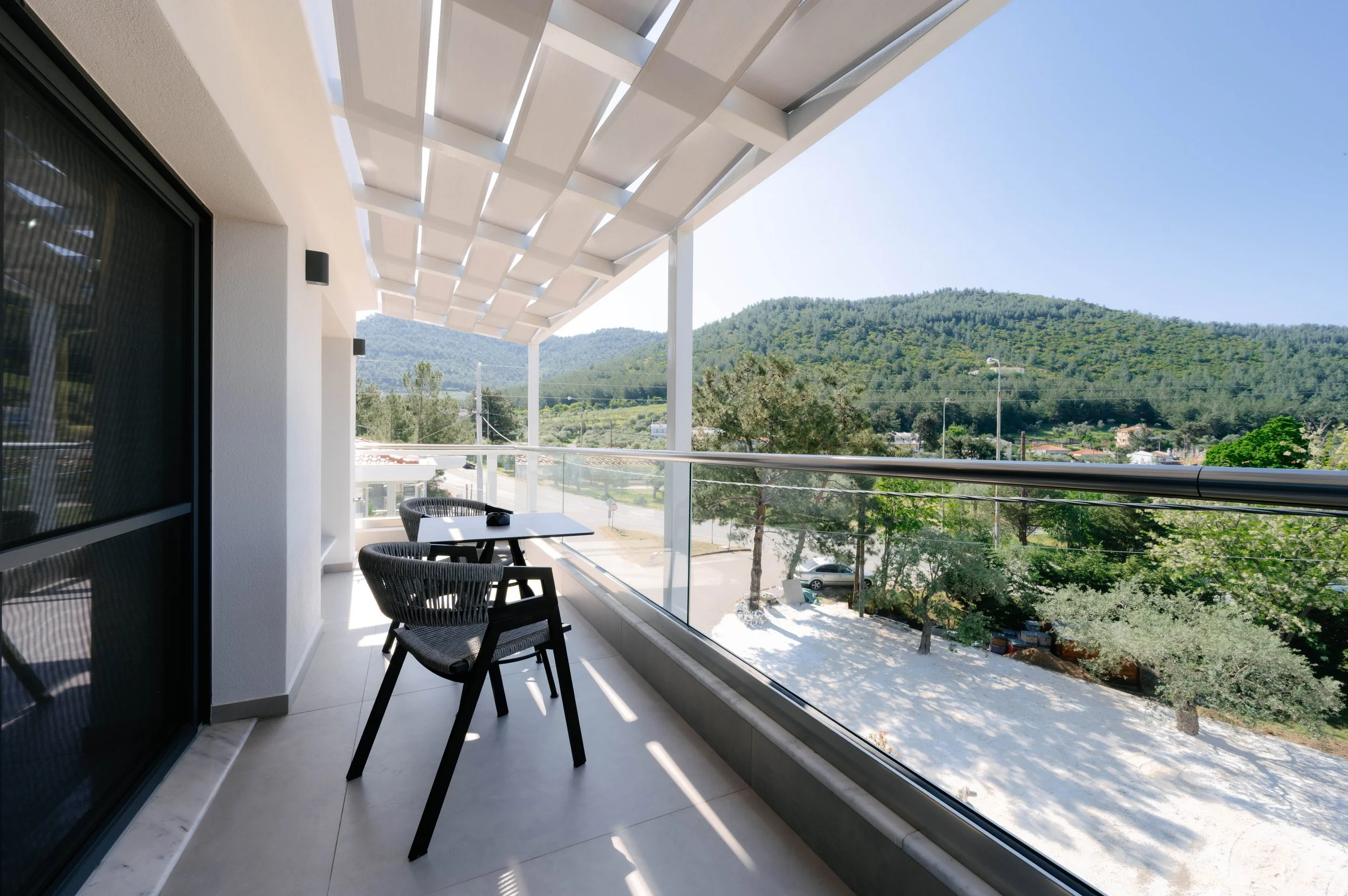 Balcony with two chairs and a table overlooking a tree-filled landscape with hills in the background under a clear blue sky.