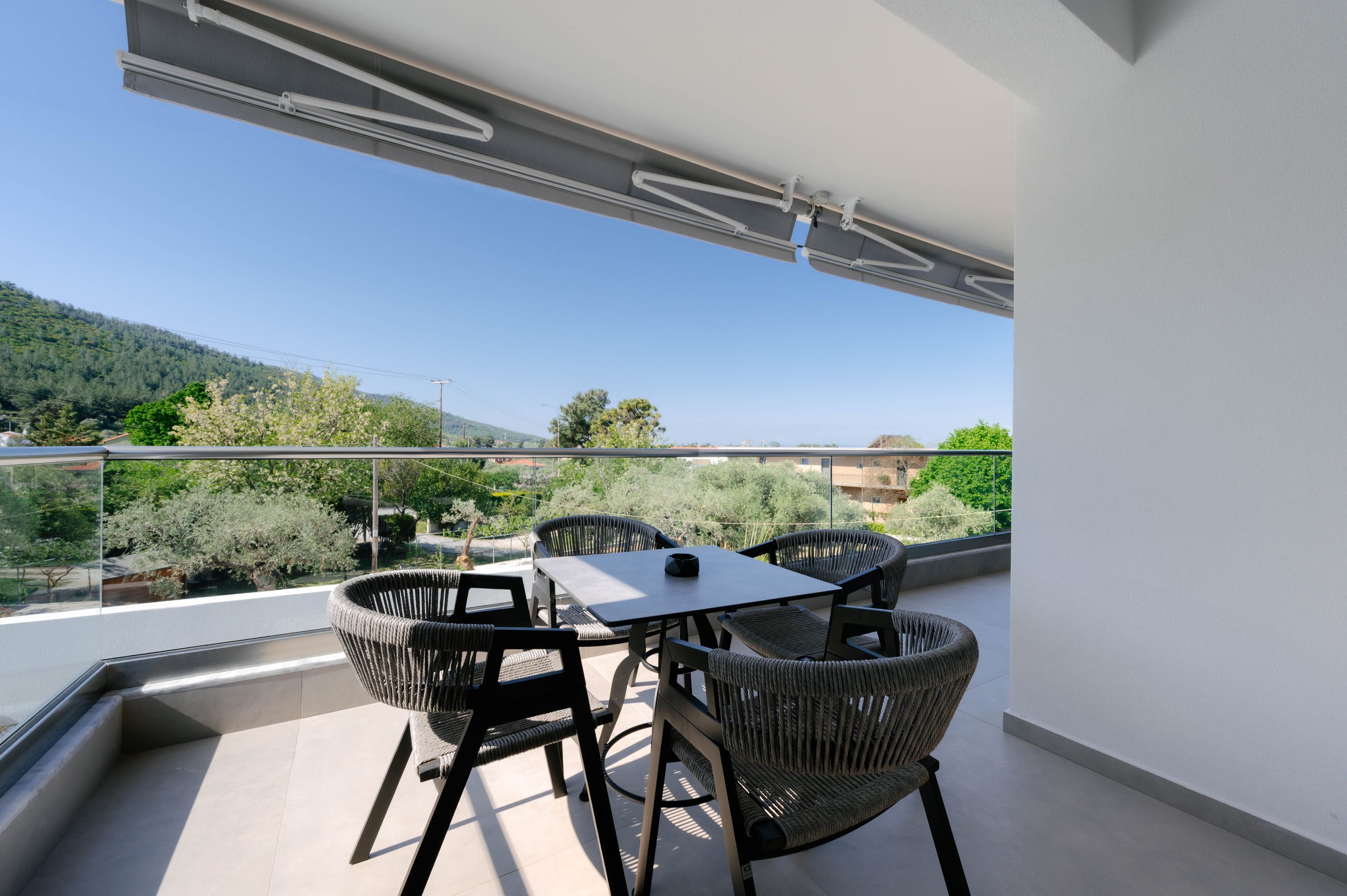 Balcony with a table and four black woven chairs, overlooking a lush green landscape with trees and hills under a clear blue sky.