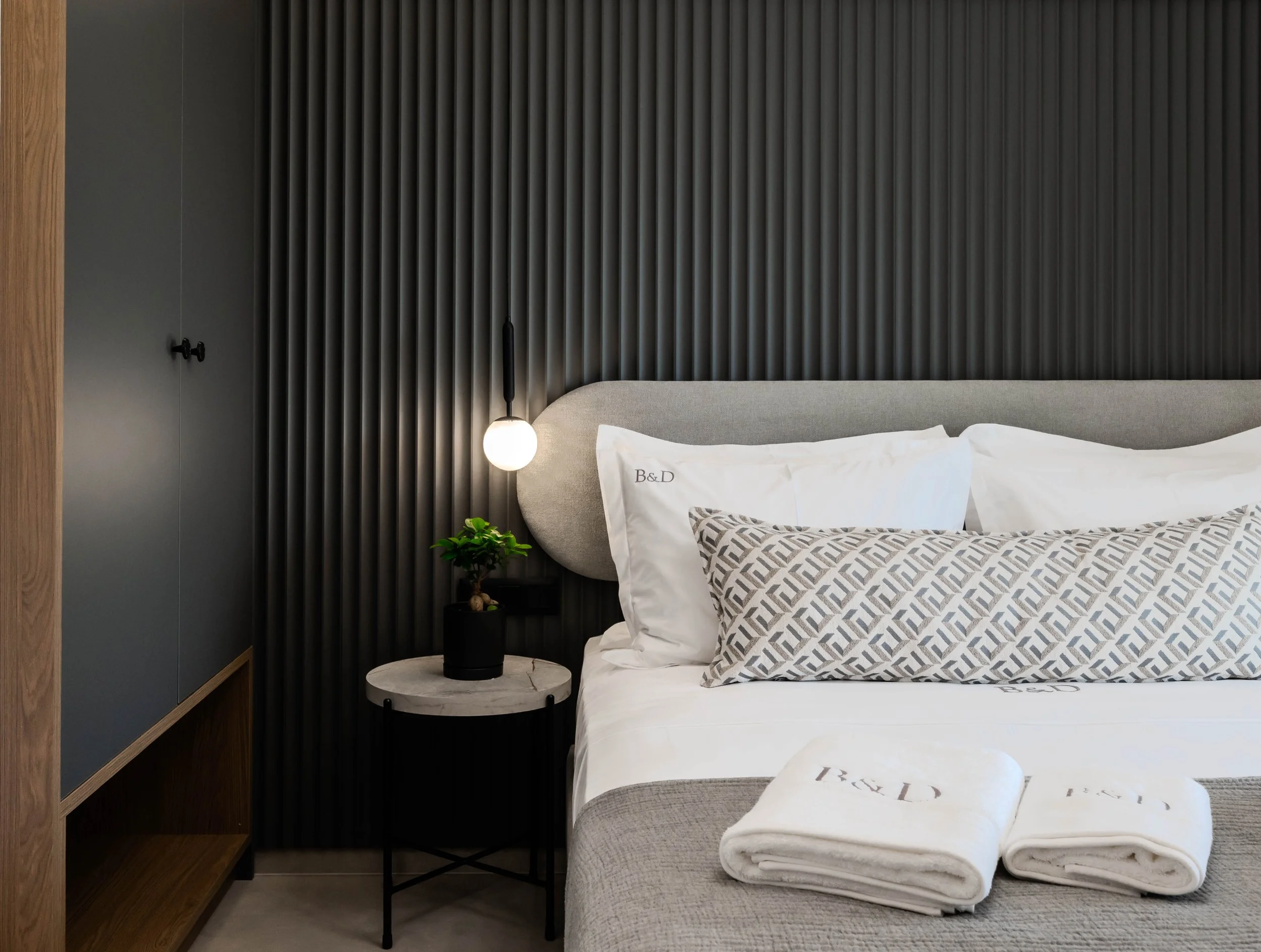A modern bedroom with a bed that has white sheets and pillows, a decorative pillow with a geometric pattern, a small side table with a potted plant, a hanging light, and a dark wall with vertical ridges.