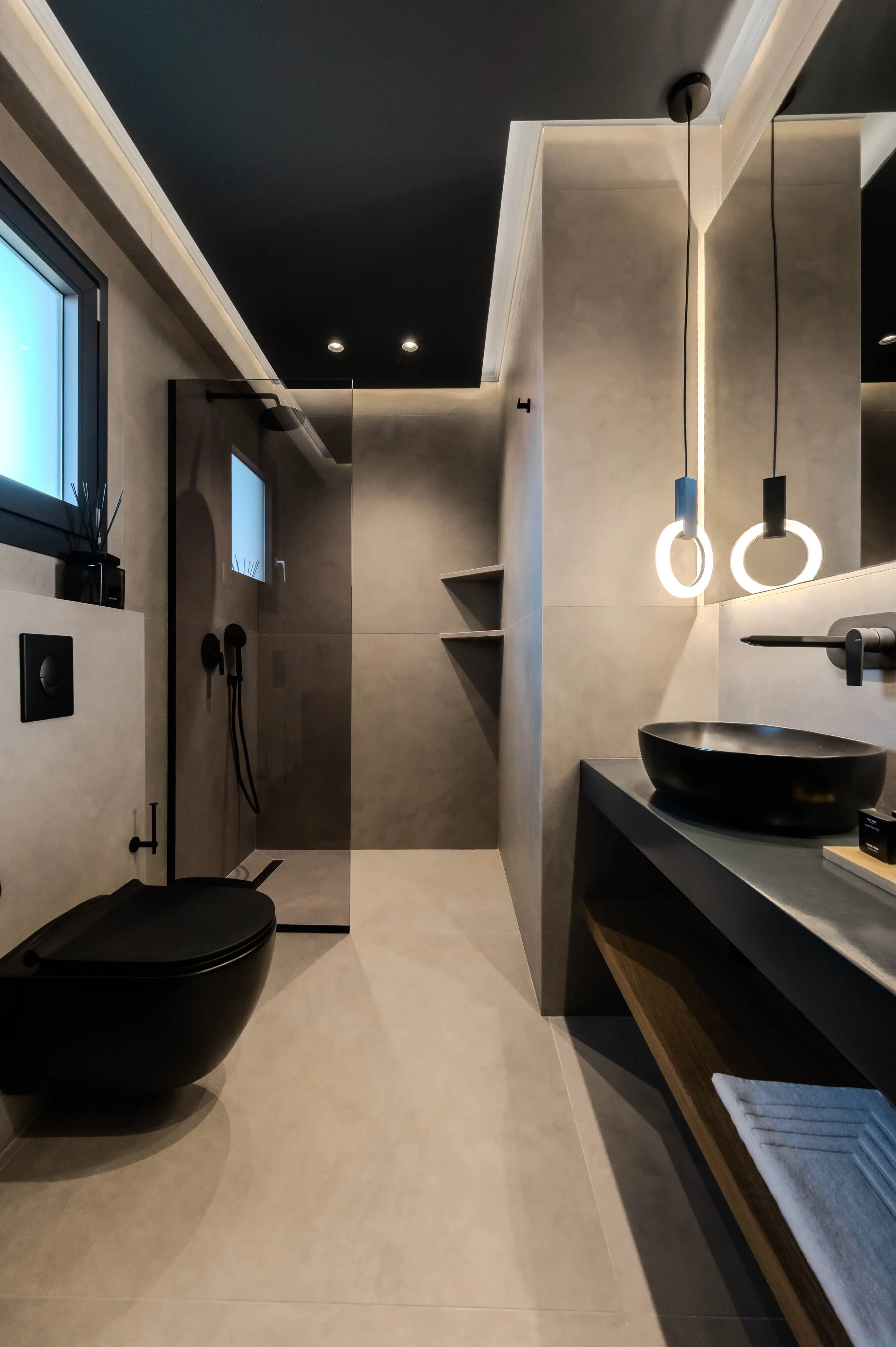 Modern bathroom with black toilet, walk-in shower with black fixtures, two hanging pendant lights, a black vessel sink on a dark countertop, and beige tiled floors and walls with some open shelving.