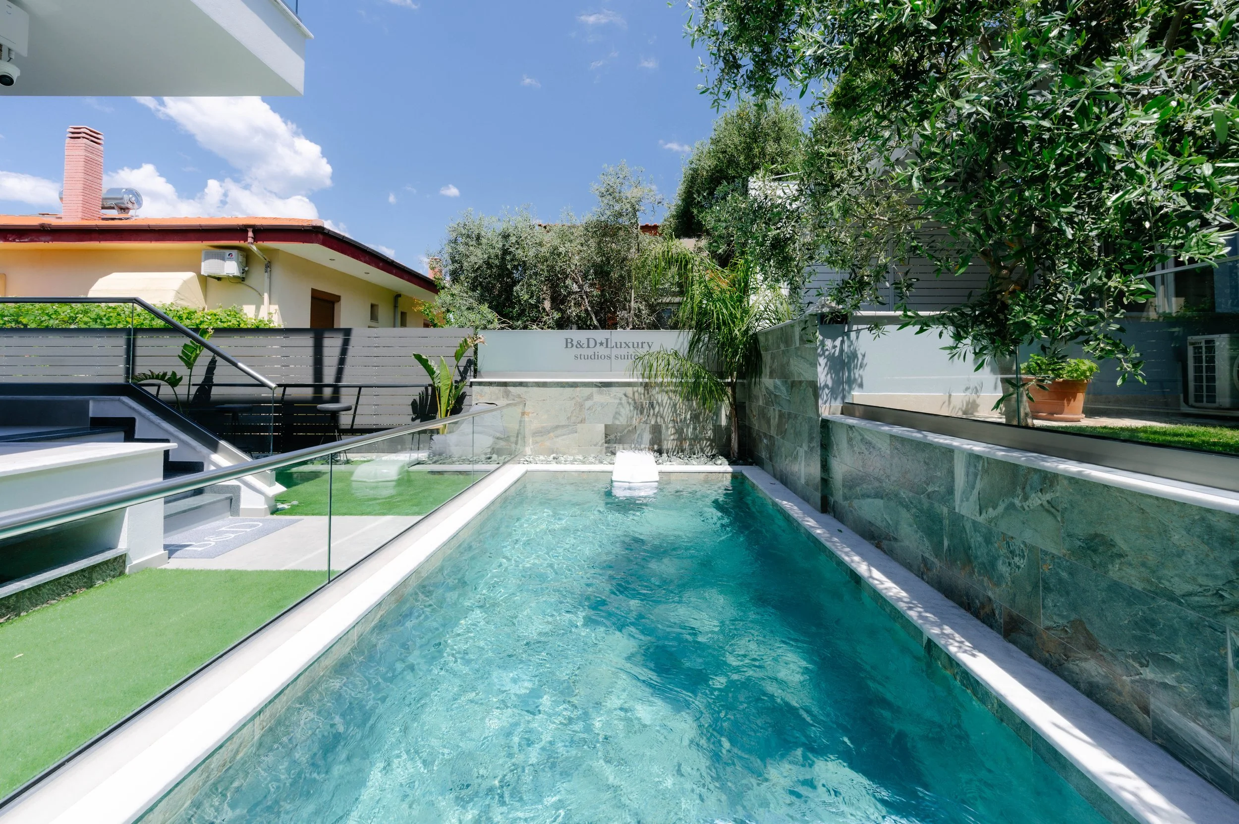 Outdoor swimming pool with glass railing, surrounded by greenery, and a sign reading 'B&D Luxury studios suite' in the background under a blue sky.