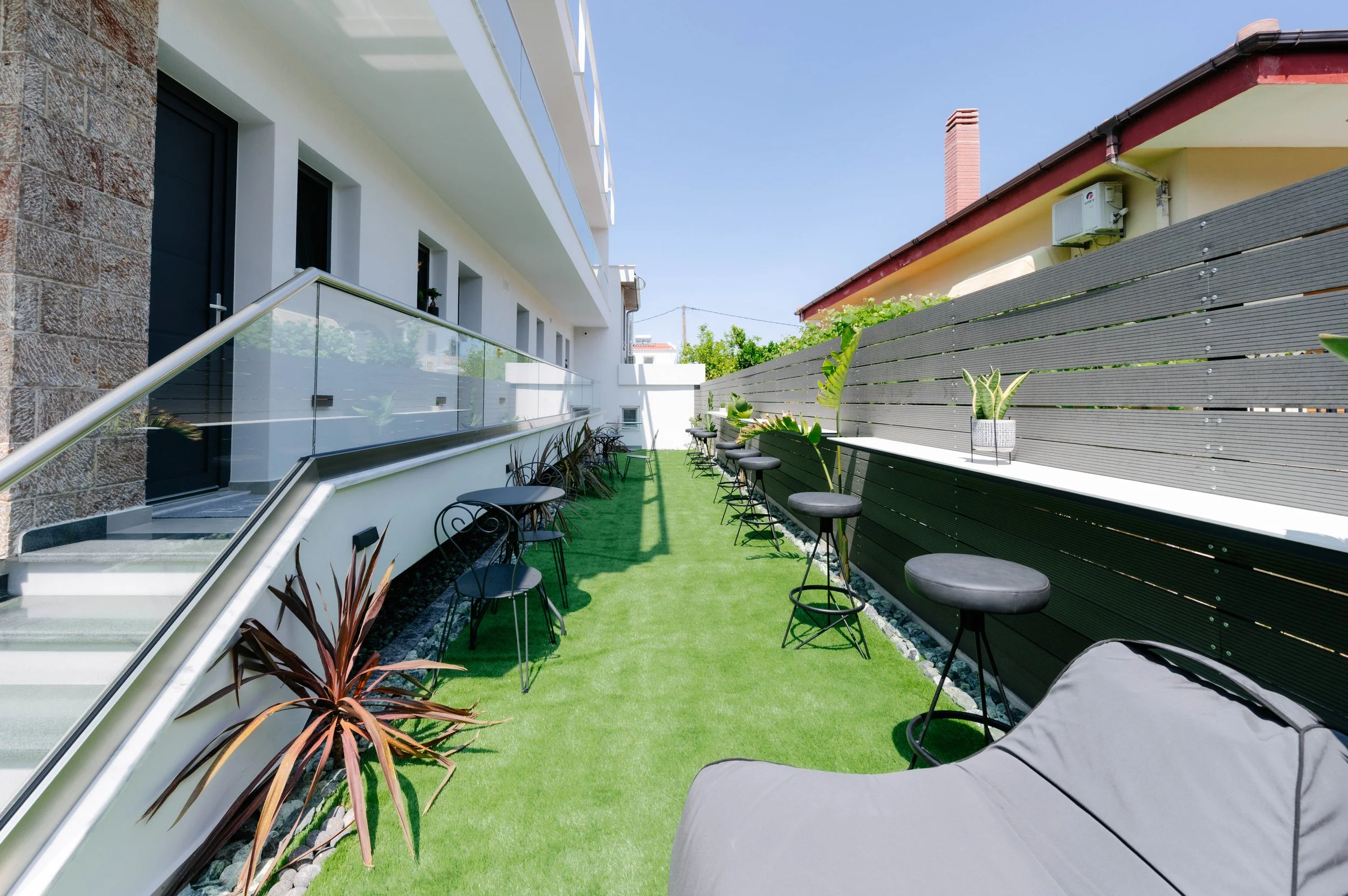Outdoor balcony area with artificial grass, black chairs, and barstools with pillows, surrounded by a black wooden fence, with potted plants on a white ledge and a gray lounge chair in the foreground, under a blue sky.
