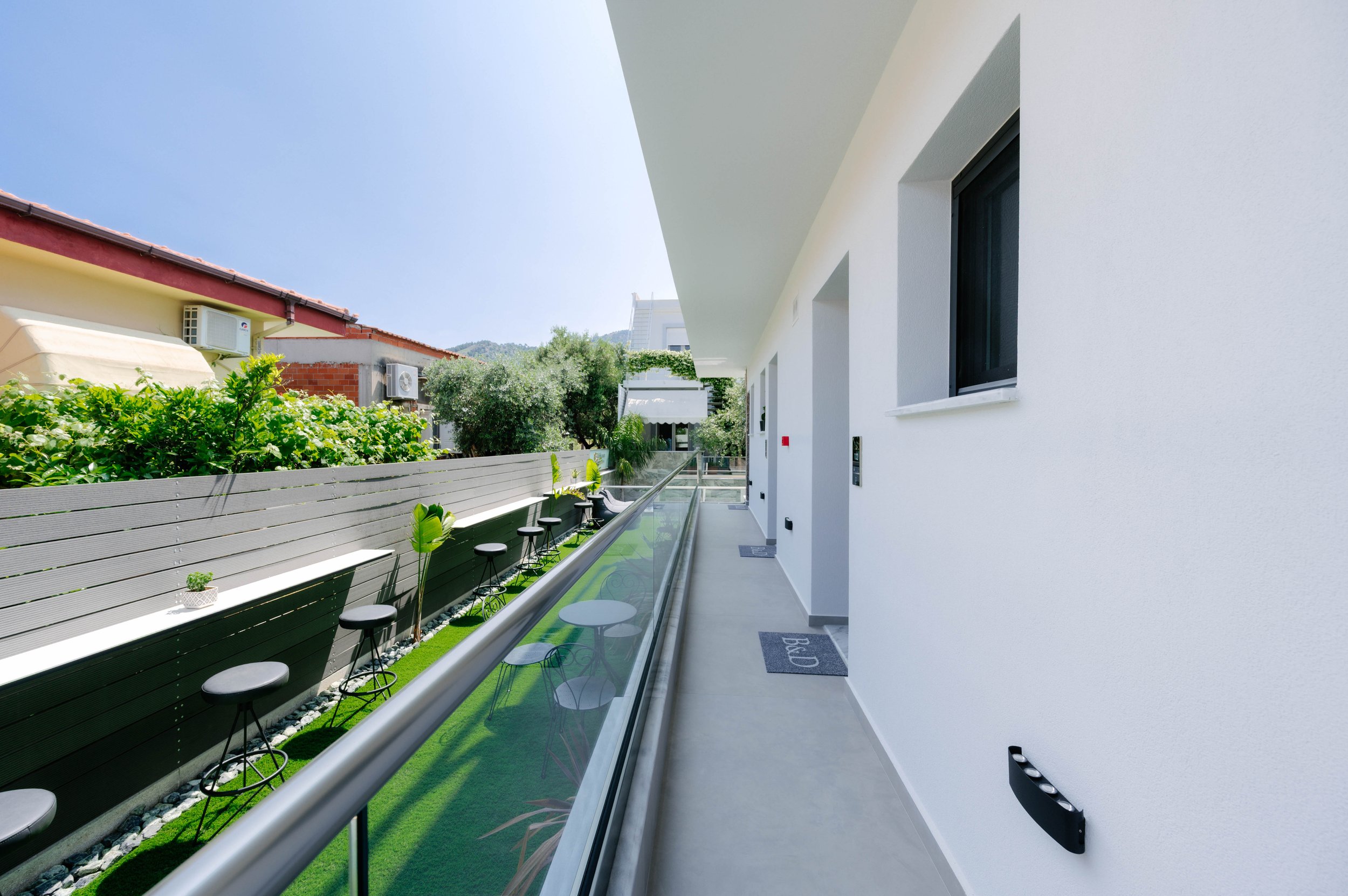 A narrow outdoor balcony with a glass railing, white walls, and potted plants, overlooking neighboring houses and greenery under a clear blue sky.