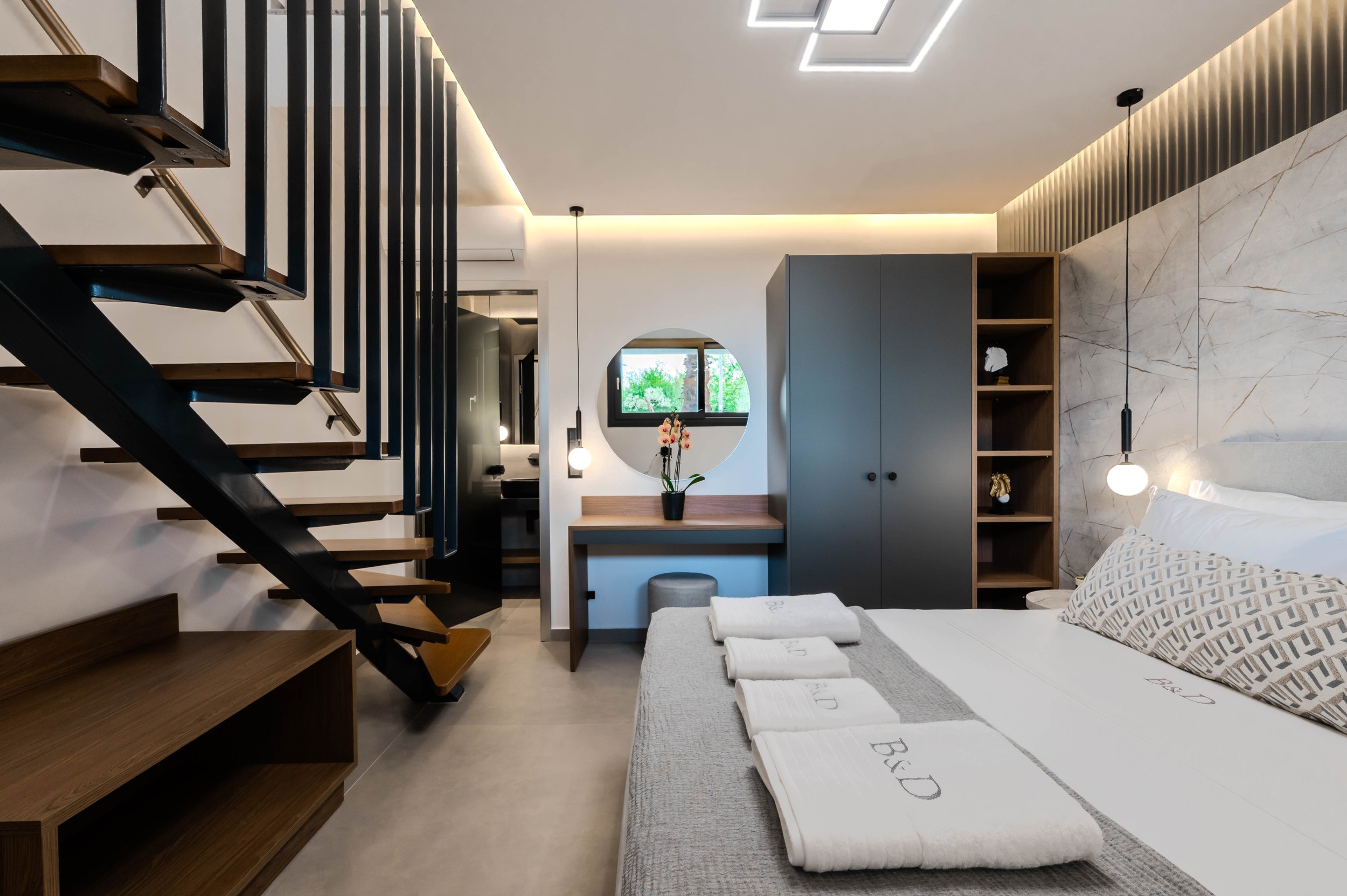 Modern hotel room with a bed, decorative pillows, and towels, a wooden staircase, a wardrobe, a vanity area with a round mirror, and contemporary lighting