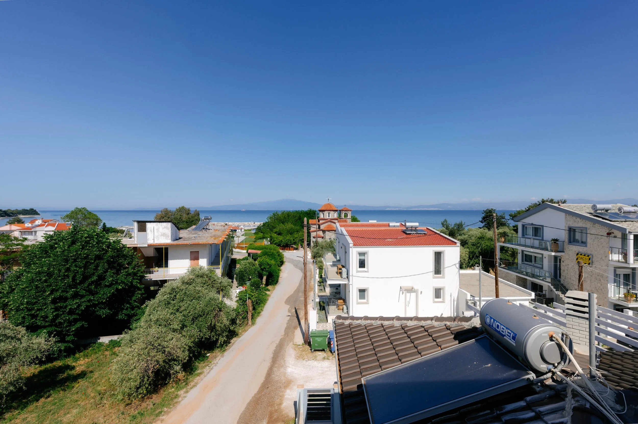 View of a coastal neighborhood with houses, trees, a dirt road, and a view of the water and mountains in the distance under a clear blue sky.