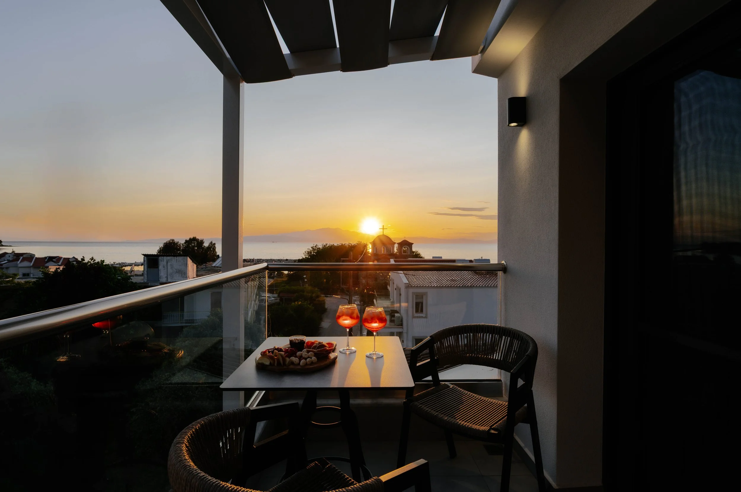 Balcony with table set with two glasses of rosé wine and a plate of snacks, overlooking the sunset over the ocean and distant mountains