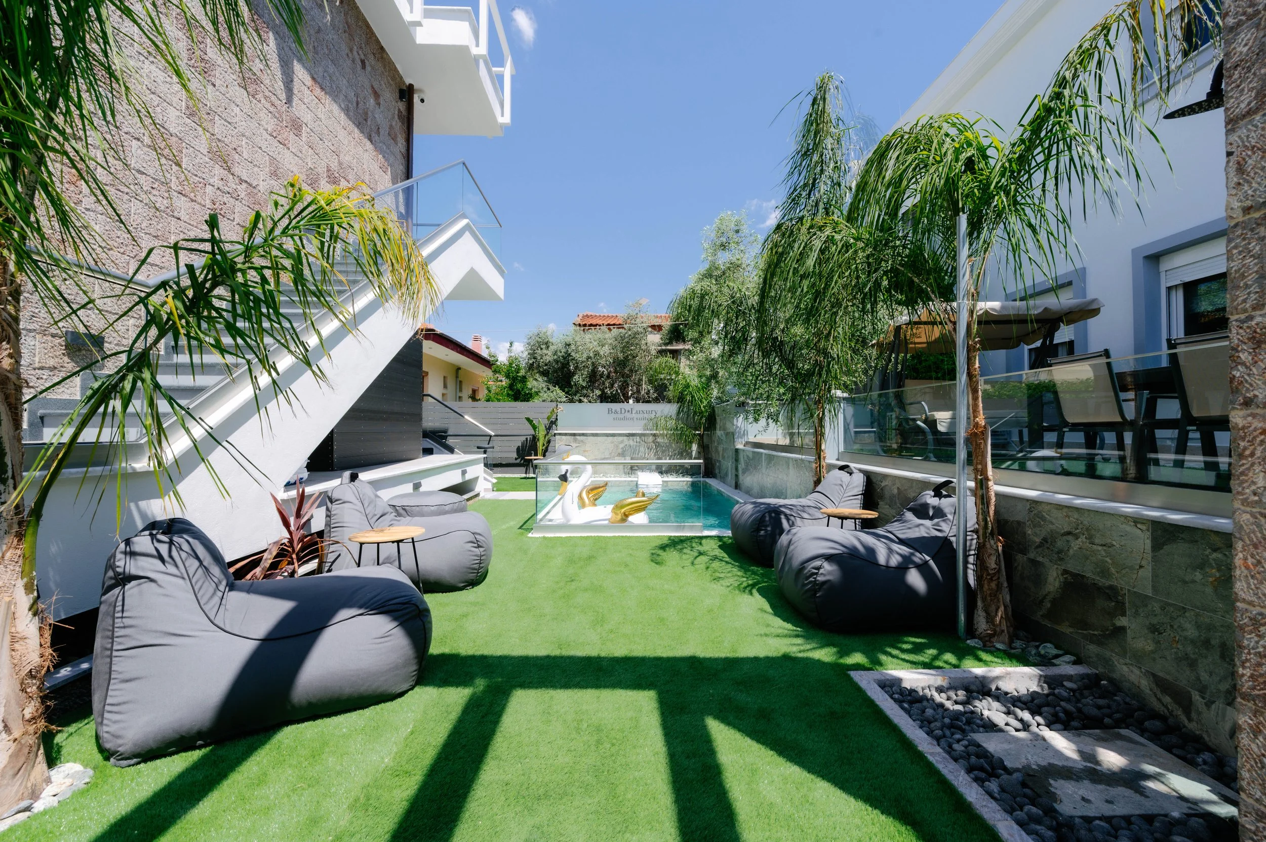 Luxury backyard with green artificial grass, black bean bag chairs, tall palms, and a small pool with floating flamingo and swan inflatables, surrounded by modern white buildings and a clear blue sky.