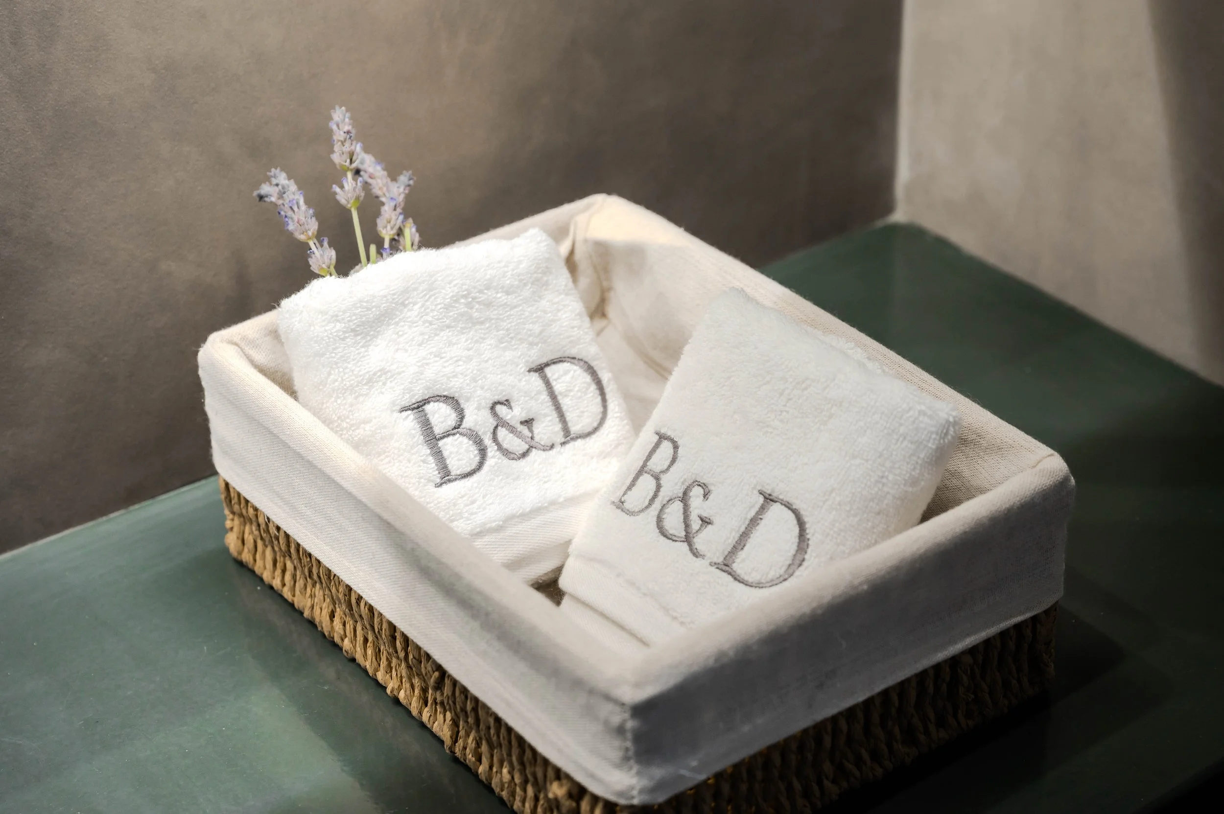 A woven basket containing two white embroidered towels with 'B&D' on them, and small lavender flowers, placed on a dark green surface.