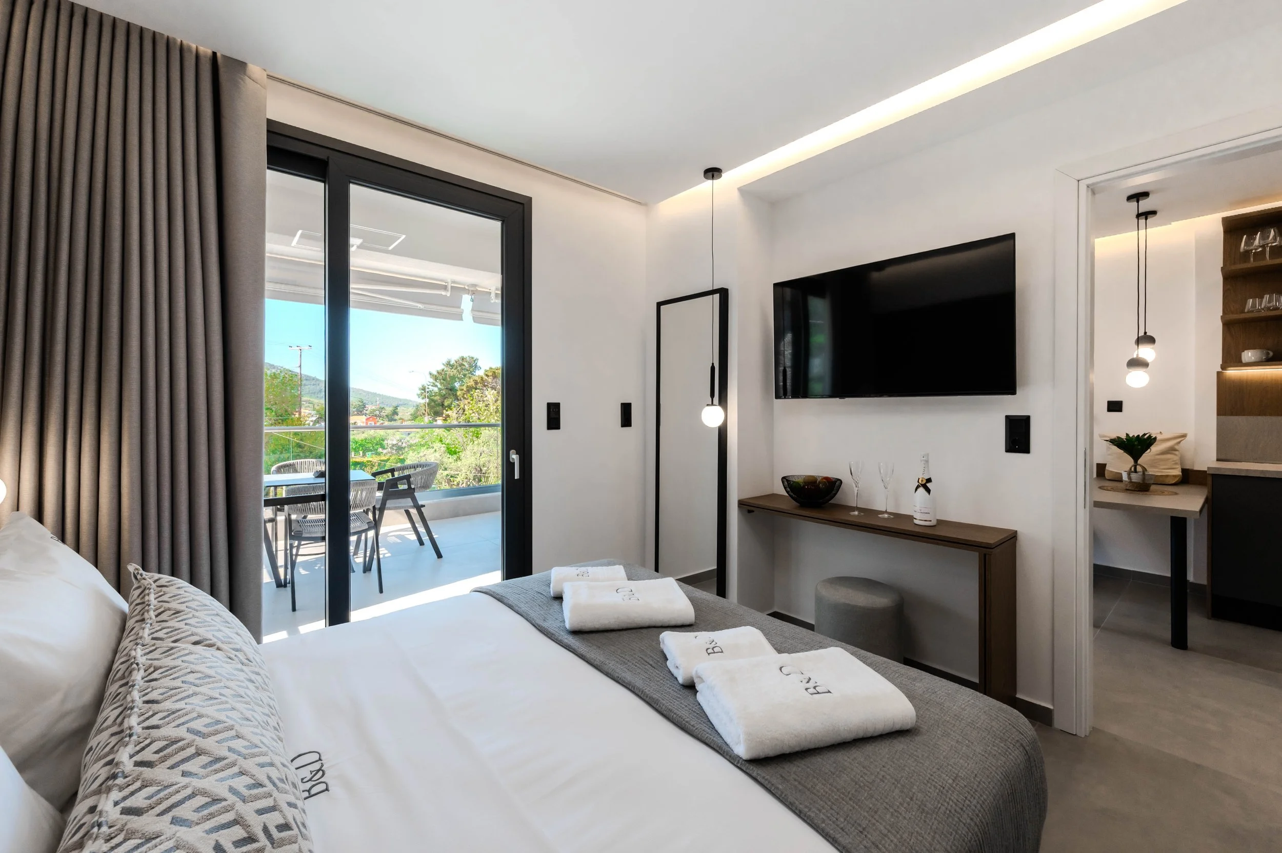 Modern hotel room with a balcony, double bed with decorative pillows and towels, wall-mounted TV, small table with wine glasses, pendant lights, and sliding glass door leading to outdoor seating with views of green trees and a blue sky.