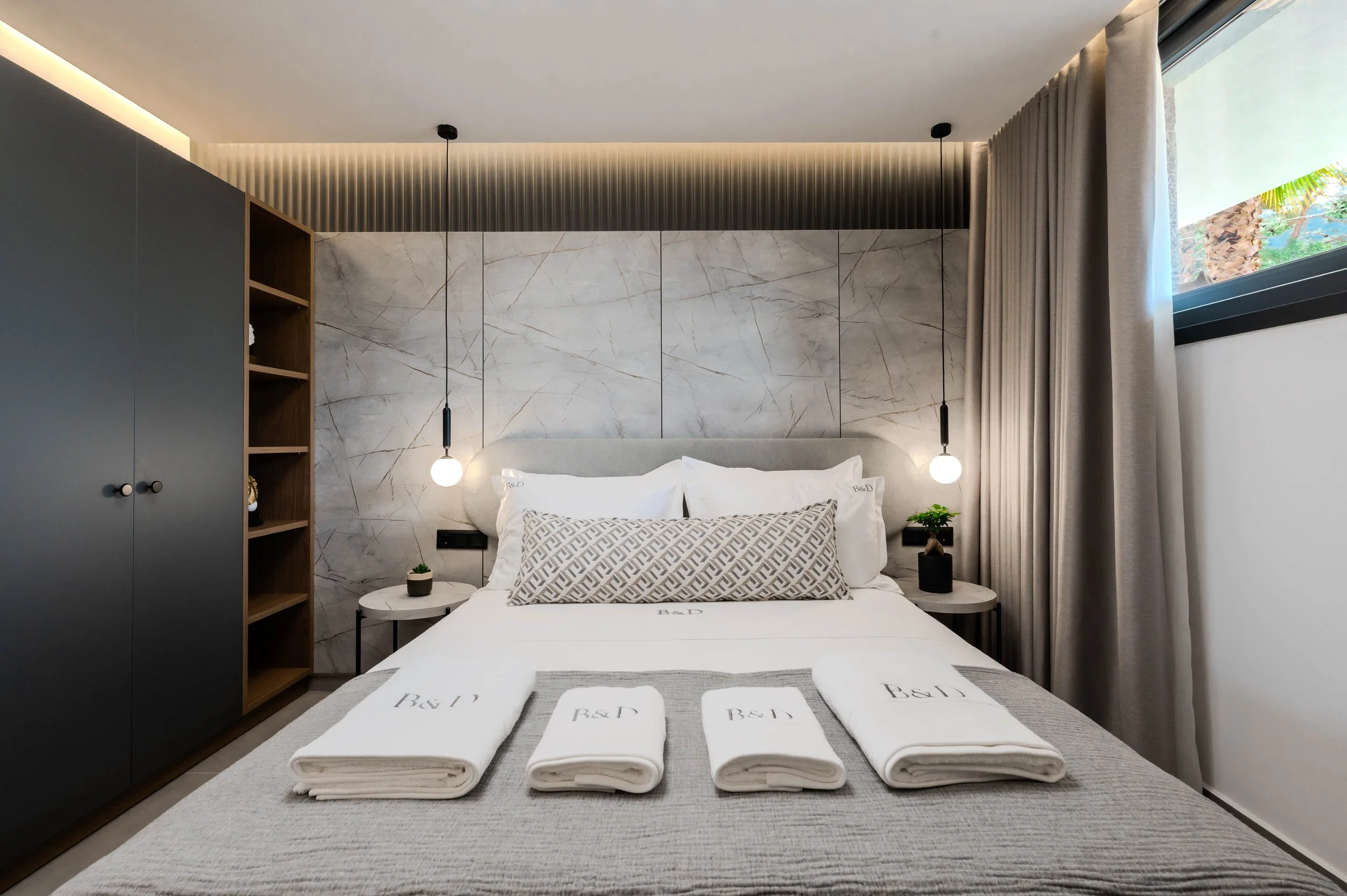 Modern bedroom with a bed covered in white linens, decorative pillows, and four rolled towels labeled 'B&J'. The headboard is against a marble-tiled wall, flanked by two side tables with small plants, and two hanging pendant lights. There's a large w