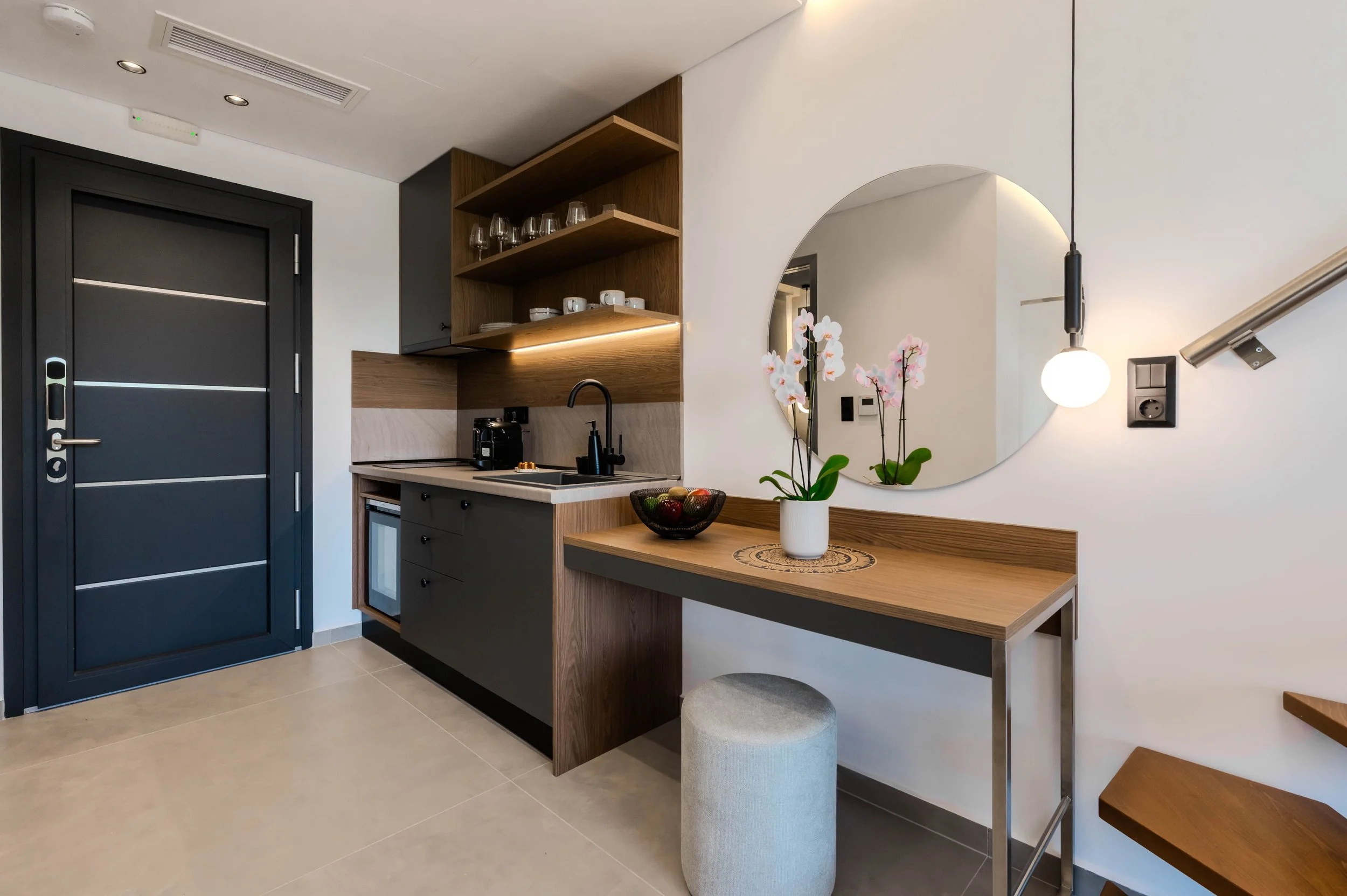 Interior of a modern small kitchen area with a black door, open wooden shelves with glasses and cups, a black countertop with a coffee machine, a white mini fridge, and a round mirror on the wall reflecting a flowery plant. There is a wooden desk wit