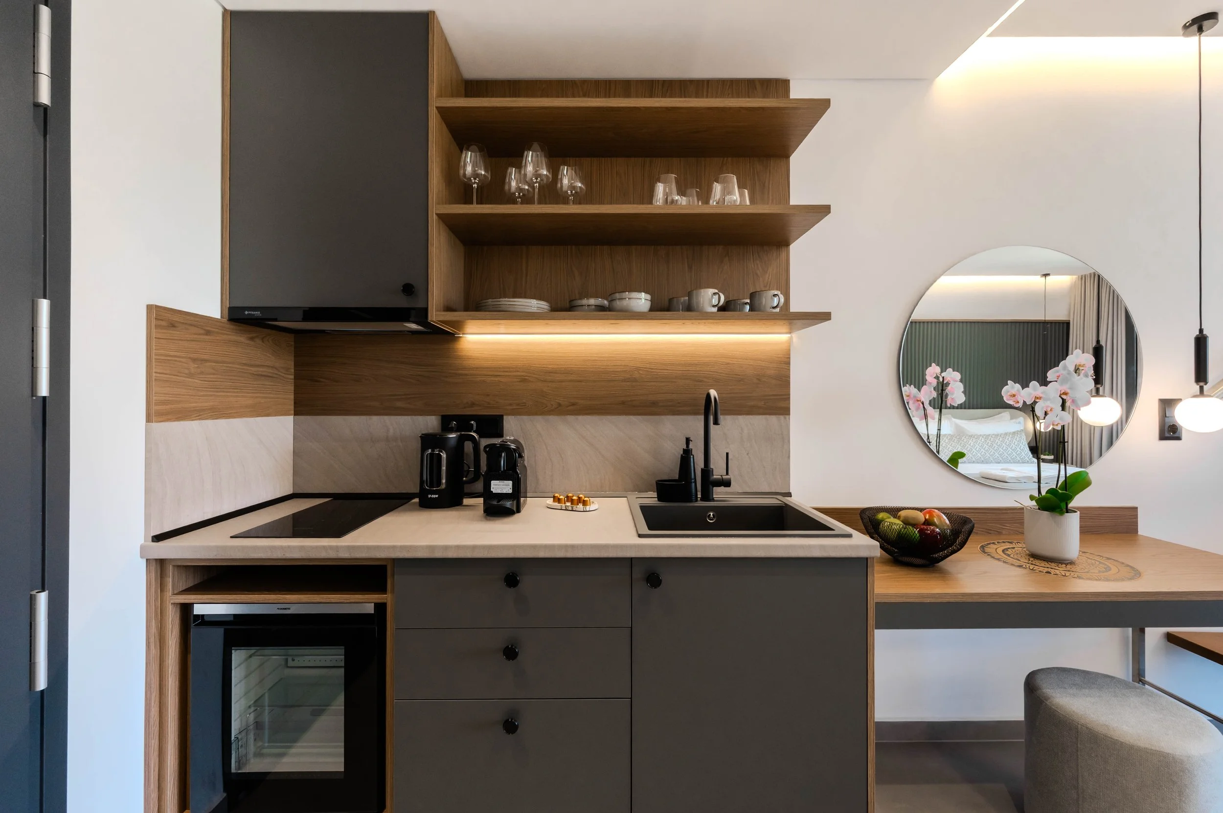 Modern kitchen with wooden shelves, black cabinets, and a small black oven. Countertop has coffee maker and kettle, with a sink nearby. A round mirror on the wall reflects a bedroom, with orchid flowers on the counter and a bowl of fruit.