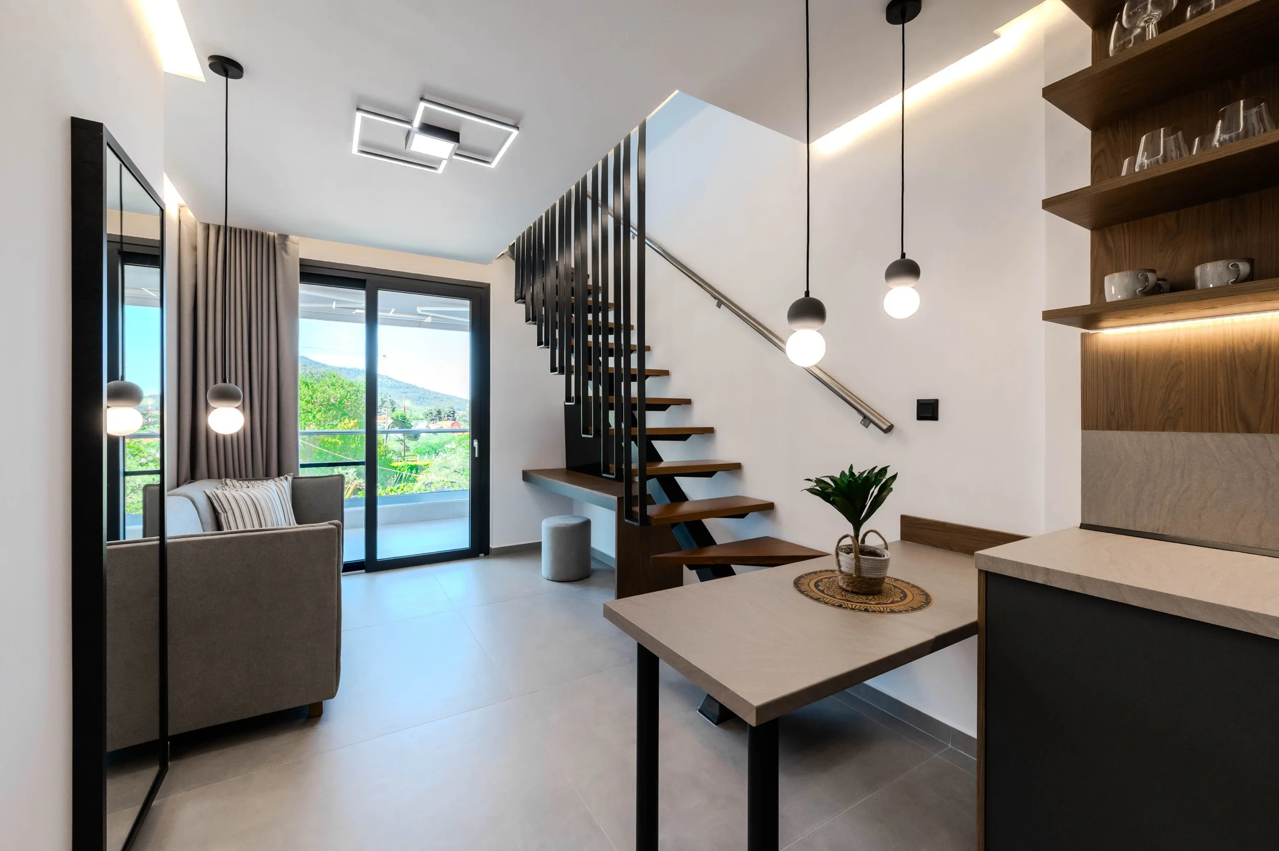 Modern living room with sliding glass door leading to balcony, staircase with black metal railing, hanging spherical pendant lights, potted plant on small table, and wooden shelves with cups and glasses.