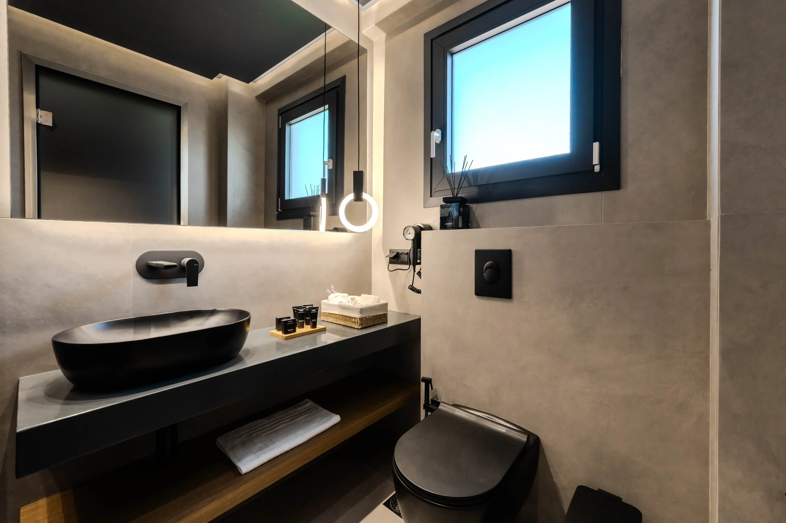 Modern bathroom with black oval sink, large mirror, two small black-framed windows, white towel underneath the sink, decorative reed diffuser, and a black toilet.