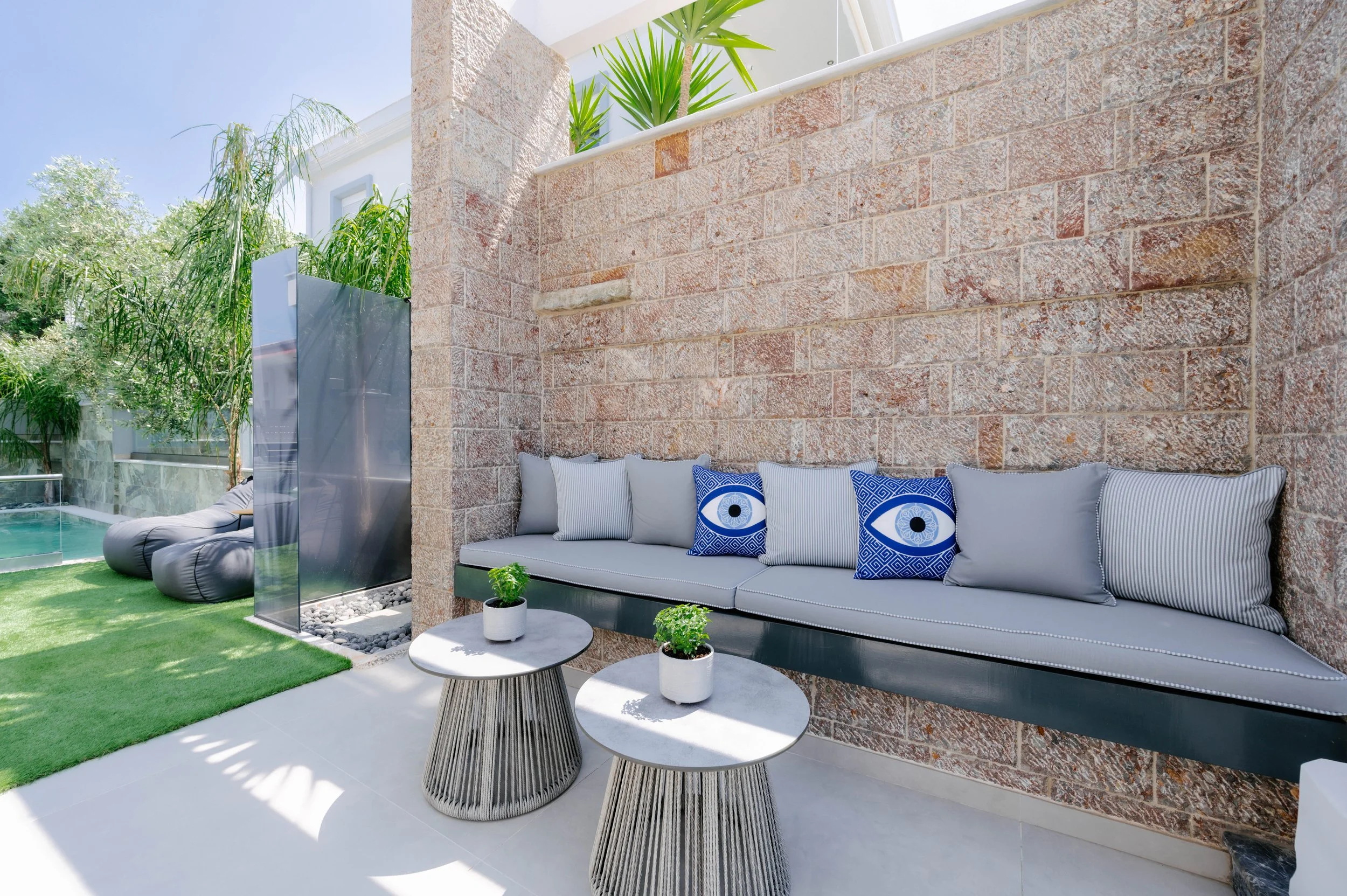 Outdoor patio area with cushioned bench against a brick wall, two small round tables with potted plants, and a lounging chair near a pool with green trees in the background.