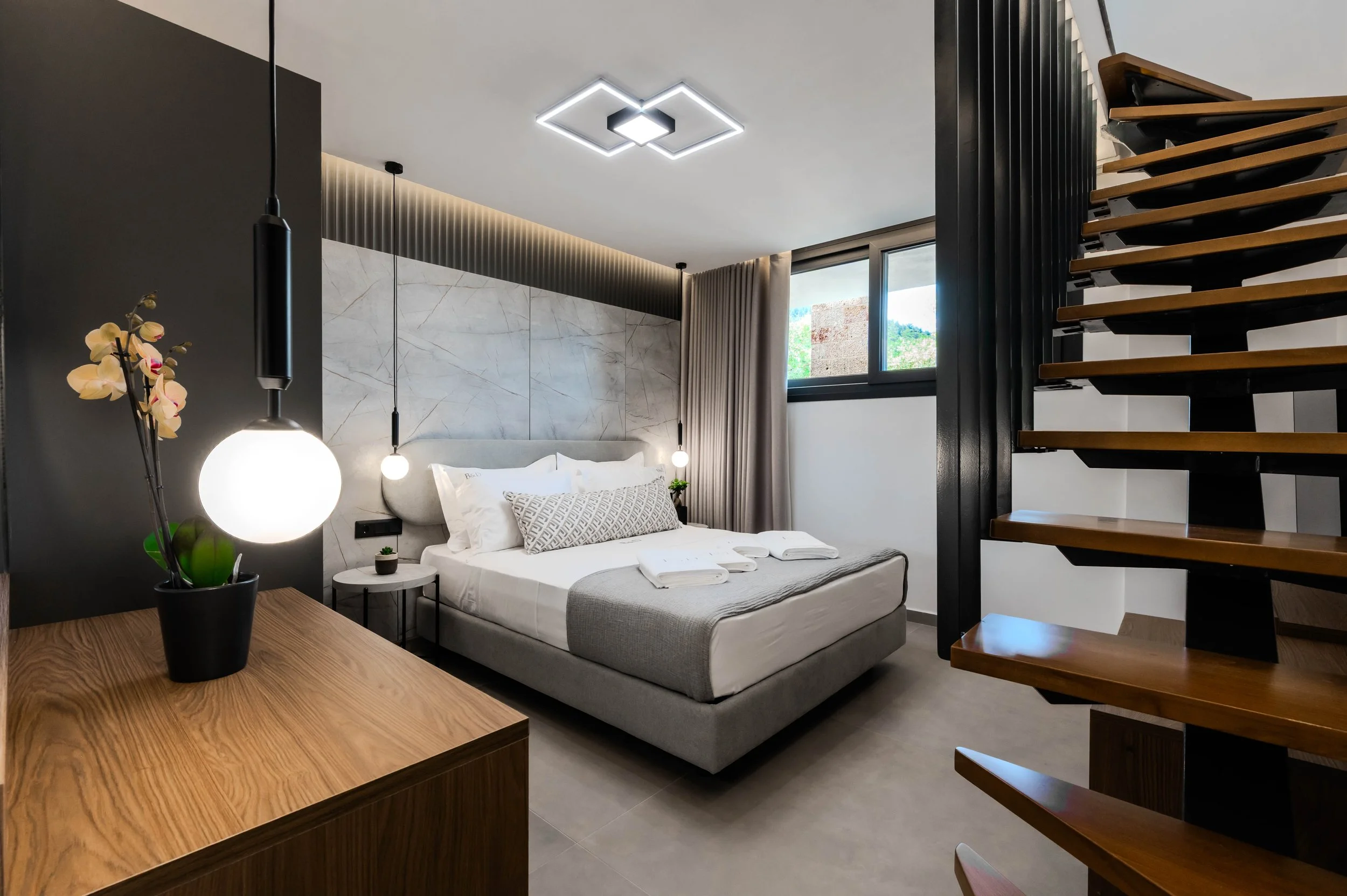 Modern bedroom with a large bed, gray and white color scheme, hanging pendant lights, a small side table, and a staircase with wooden steps leading upwards.