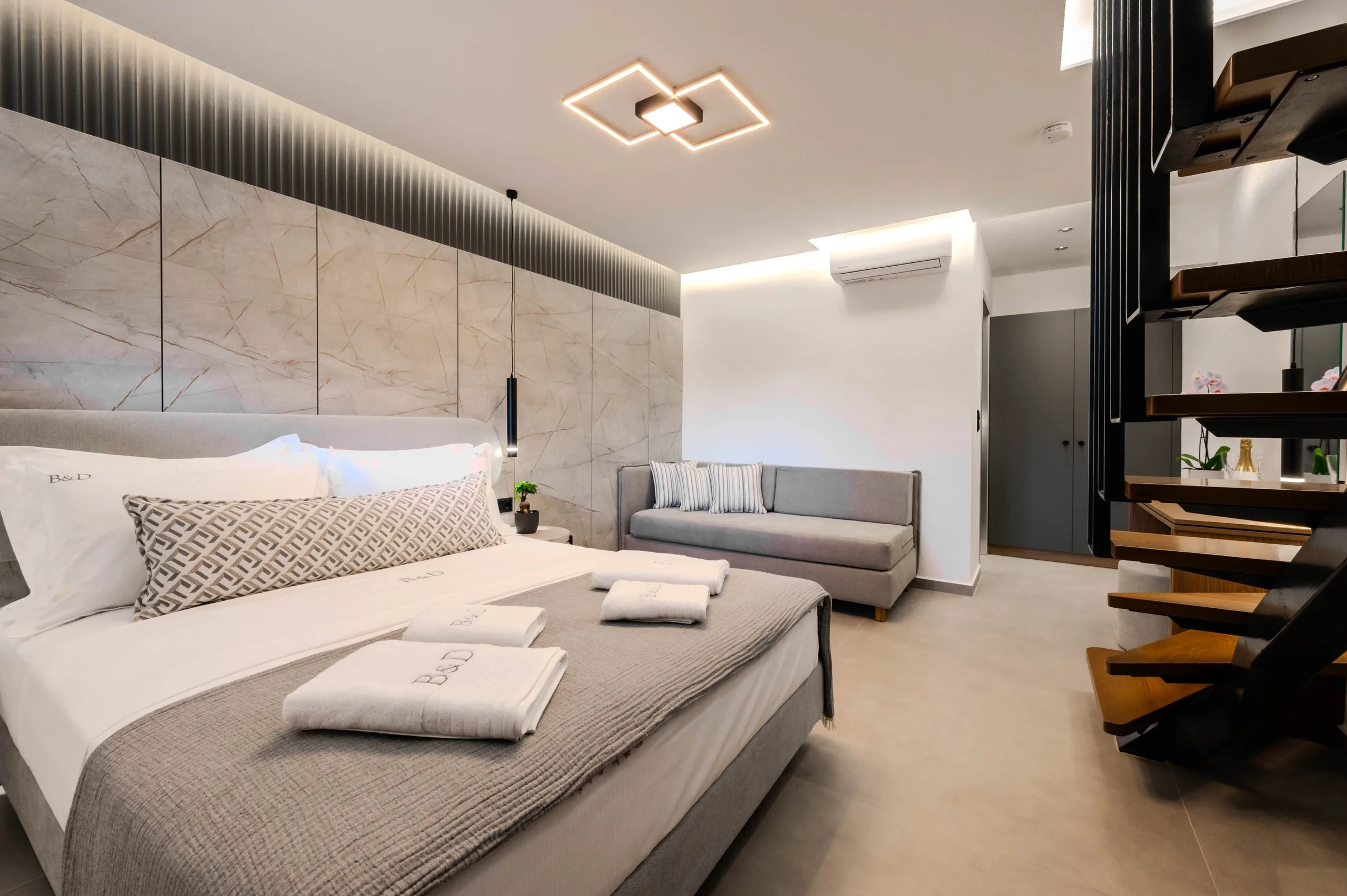 Modern hotel bedroom with a large bed, a gray sofa with striped pillows, and stylish lighting fixtures.