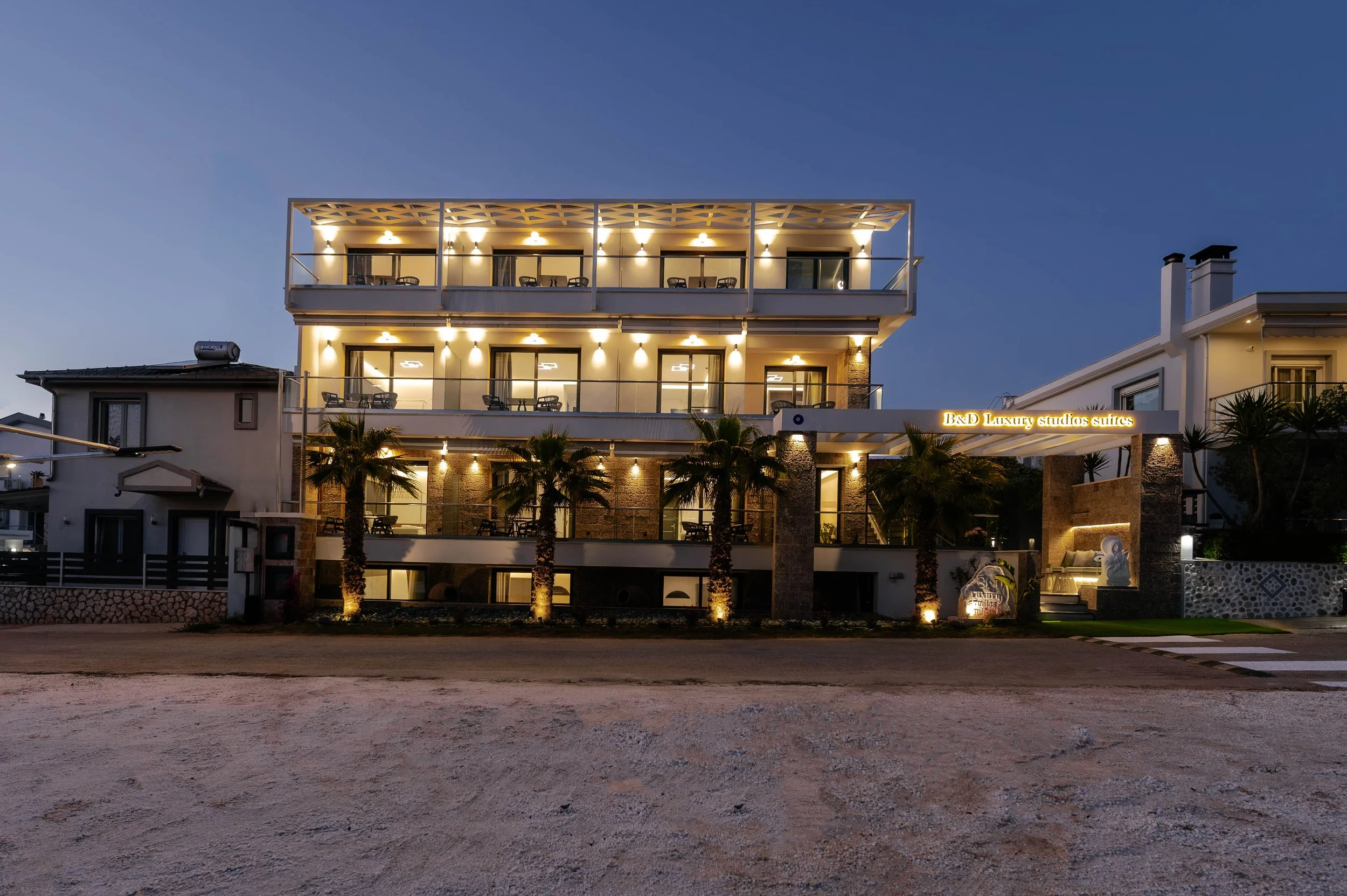 Night view of a modern hotel with three floors, illuminated from inside, featuring a sign that reads 'B&D Luxury Studios Suites,' surrounded by palm trees and neighboring buildings.