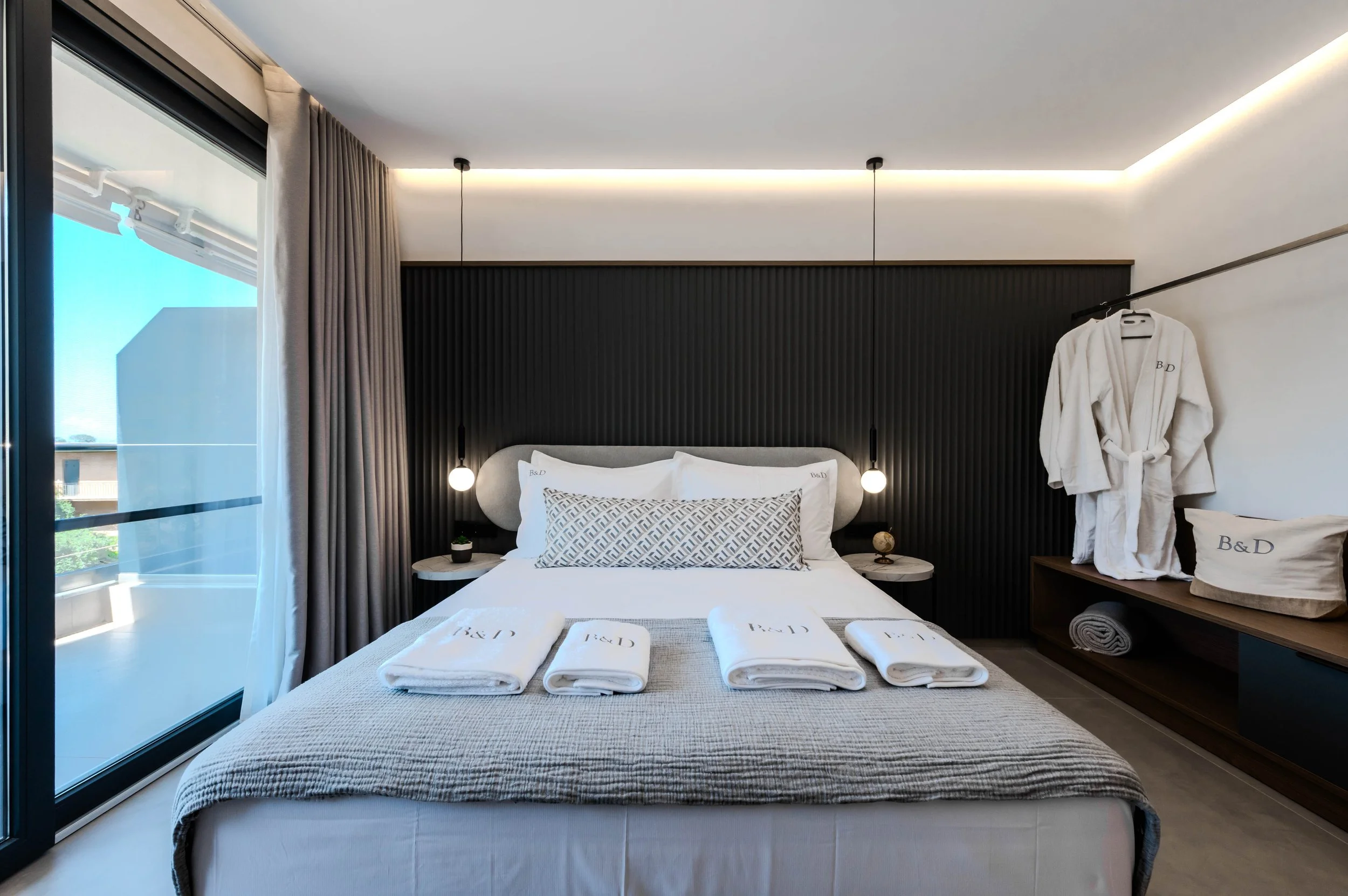 Modern hotel bedroom with a neatly made bed, towels, and robes, featuring a sliding glass door with curtains and black accent wall behind the headboard.