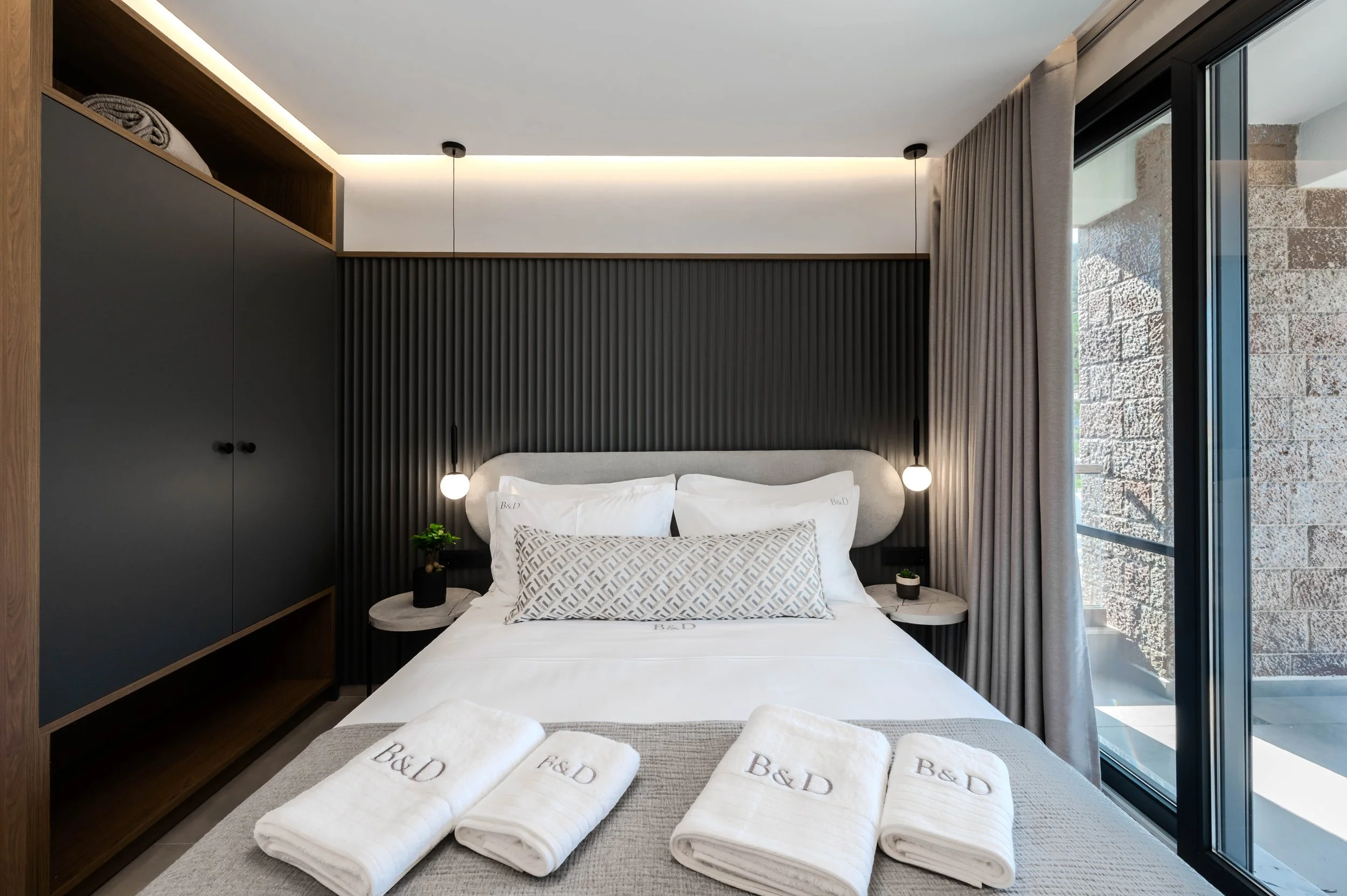 Modern hotel bedroom featuring a bed with white linens, black and white geometric pillows, bedside tables with small green plants, dark textured wall behind the bed, bedside pendant lights, large window with curtains, and neatly folded white towels o