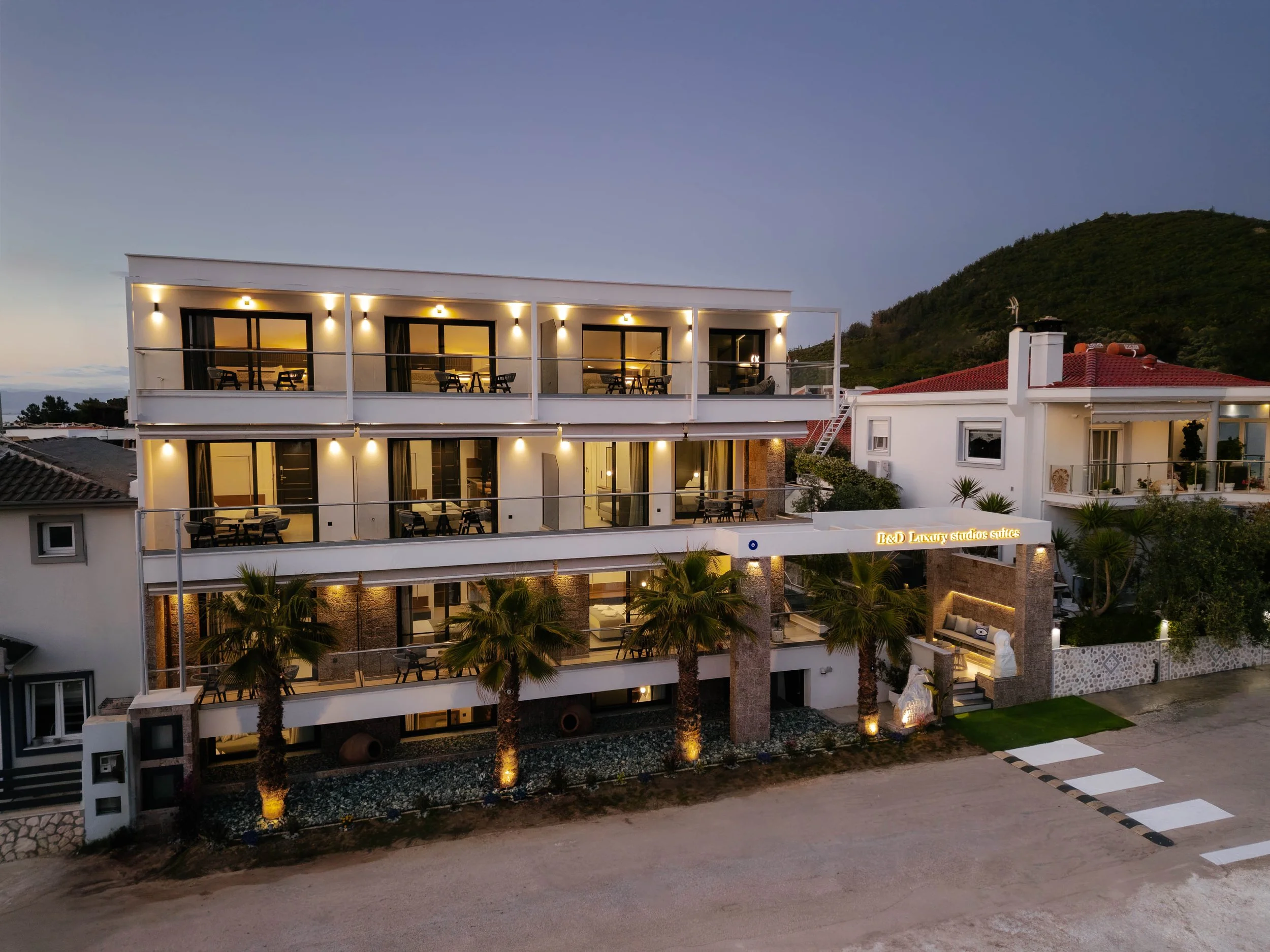 A modern, multi-story hotel or apartment building at dusk with outdoor balconies, palm trees, and illuminated exterior lights. The building has a sign that reads 'B&D Luxury studios suites'.
