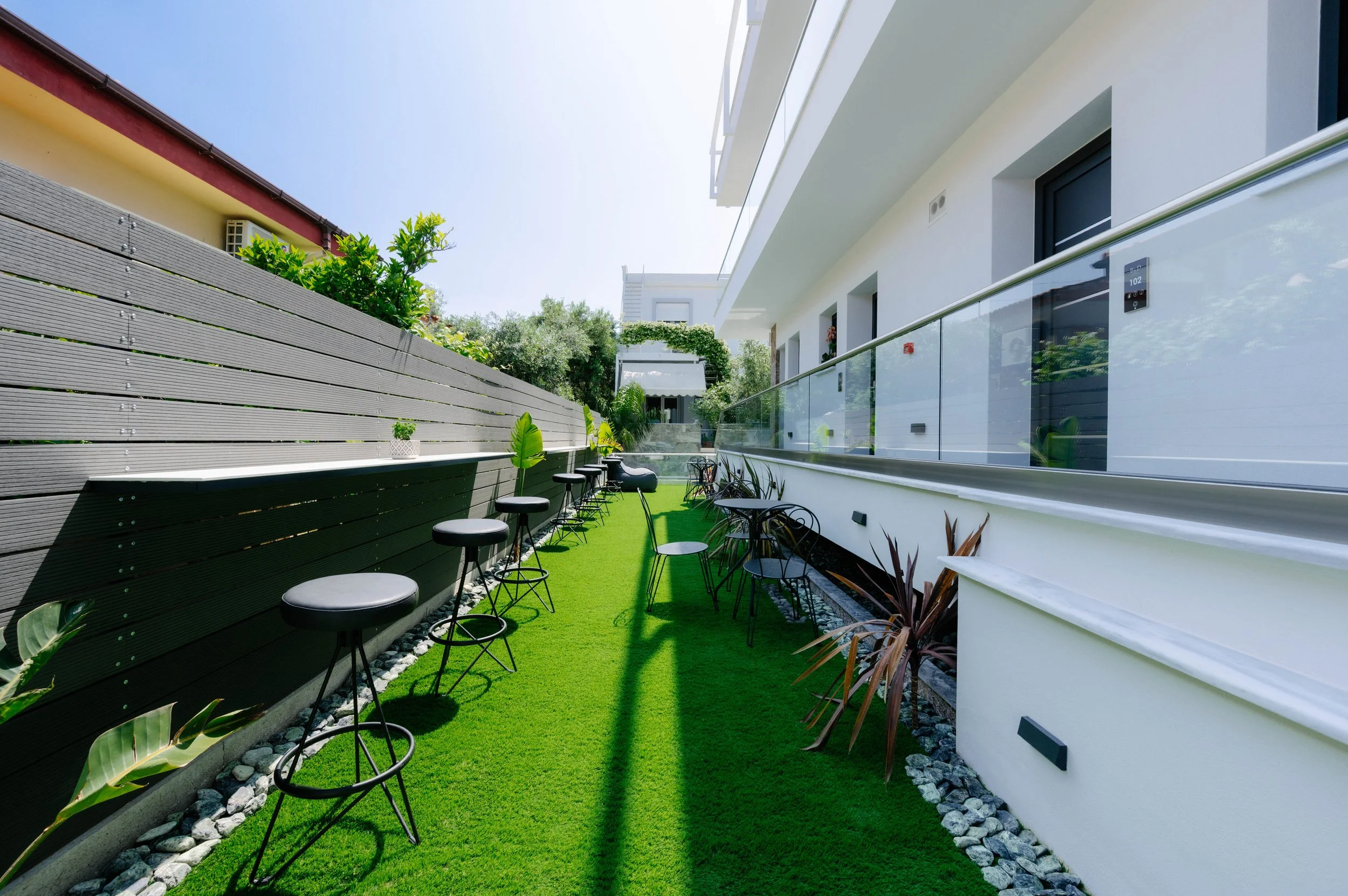 Outdoor patio with black bar stools and chairs along a green artificial grass strip, modern white building with glass railing, plants, and trees in the background under a clear blue sky.