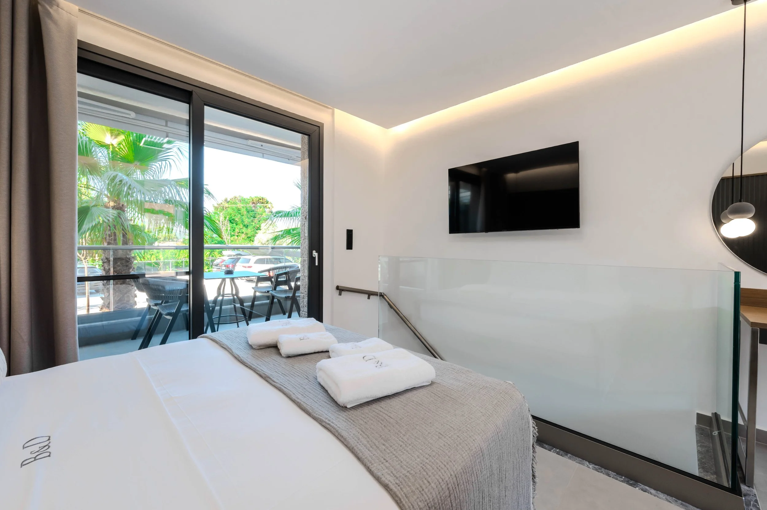 A modern hotel room with a bed, a balcony with chairs and a table, a wall-mounted TV, and tropical plants visible outside.