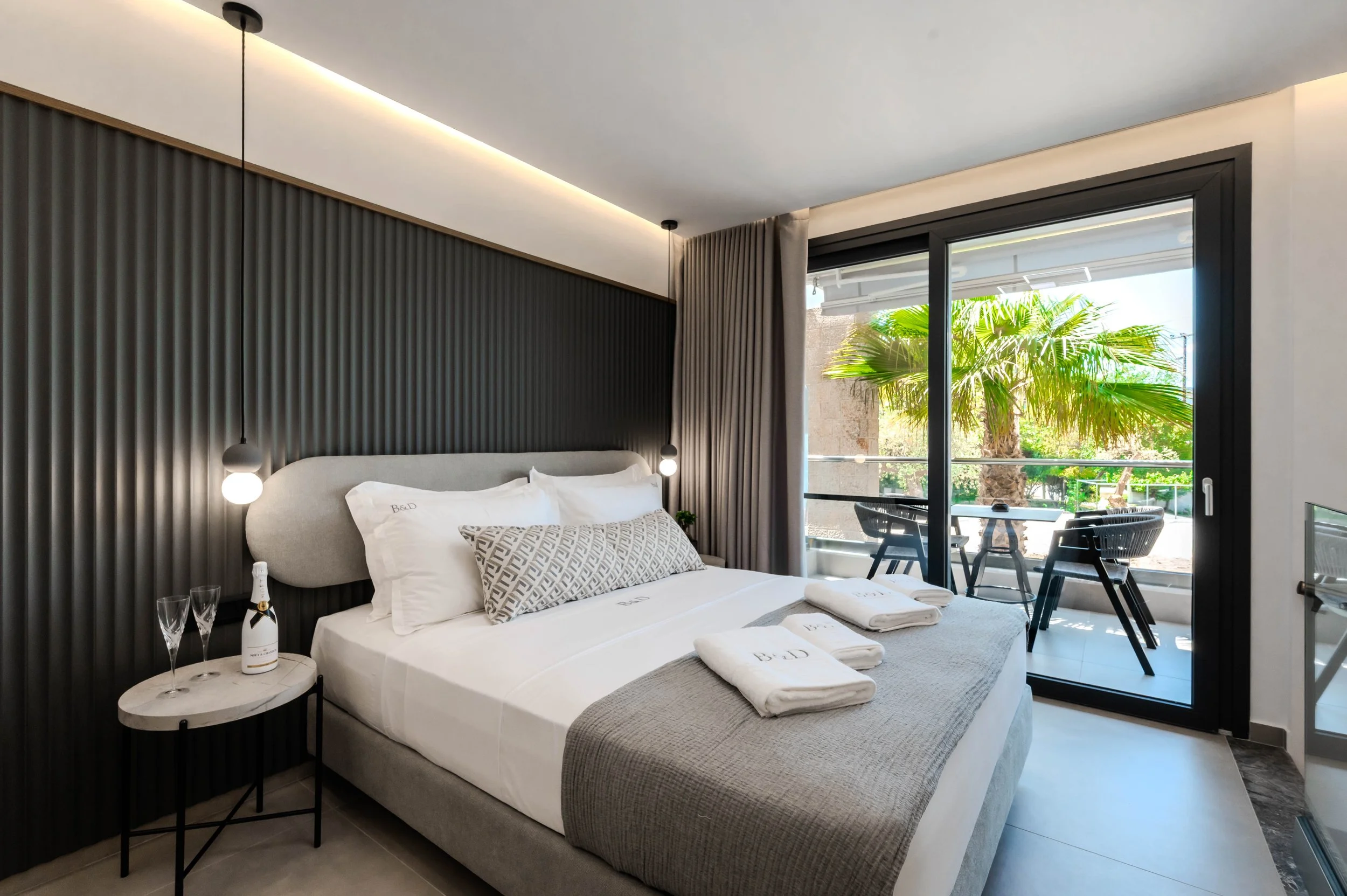 Modern bedroom with a large bed, black textured accent wall, small round side table with champagne and glasses, balcony with outdoor seating and palm trees outside.