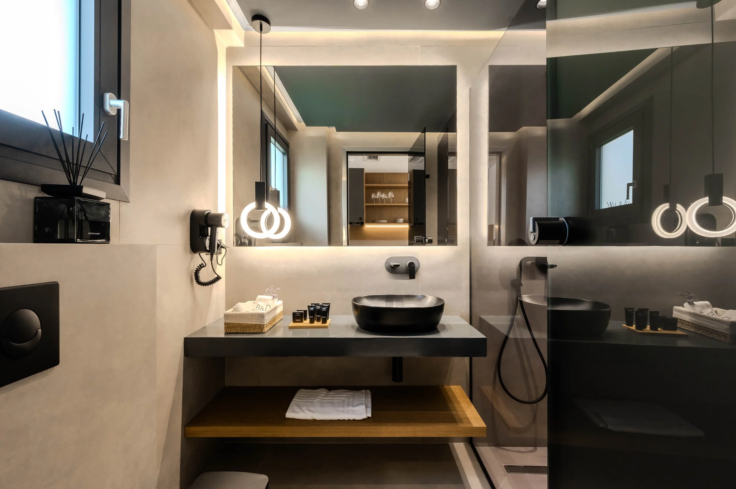 Modern bathroom with a black vessel sink, illuminated mirror, wall-mounted hairdryer, and toiletries on the gray countertop, with a shower area enclosed by glass.