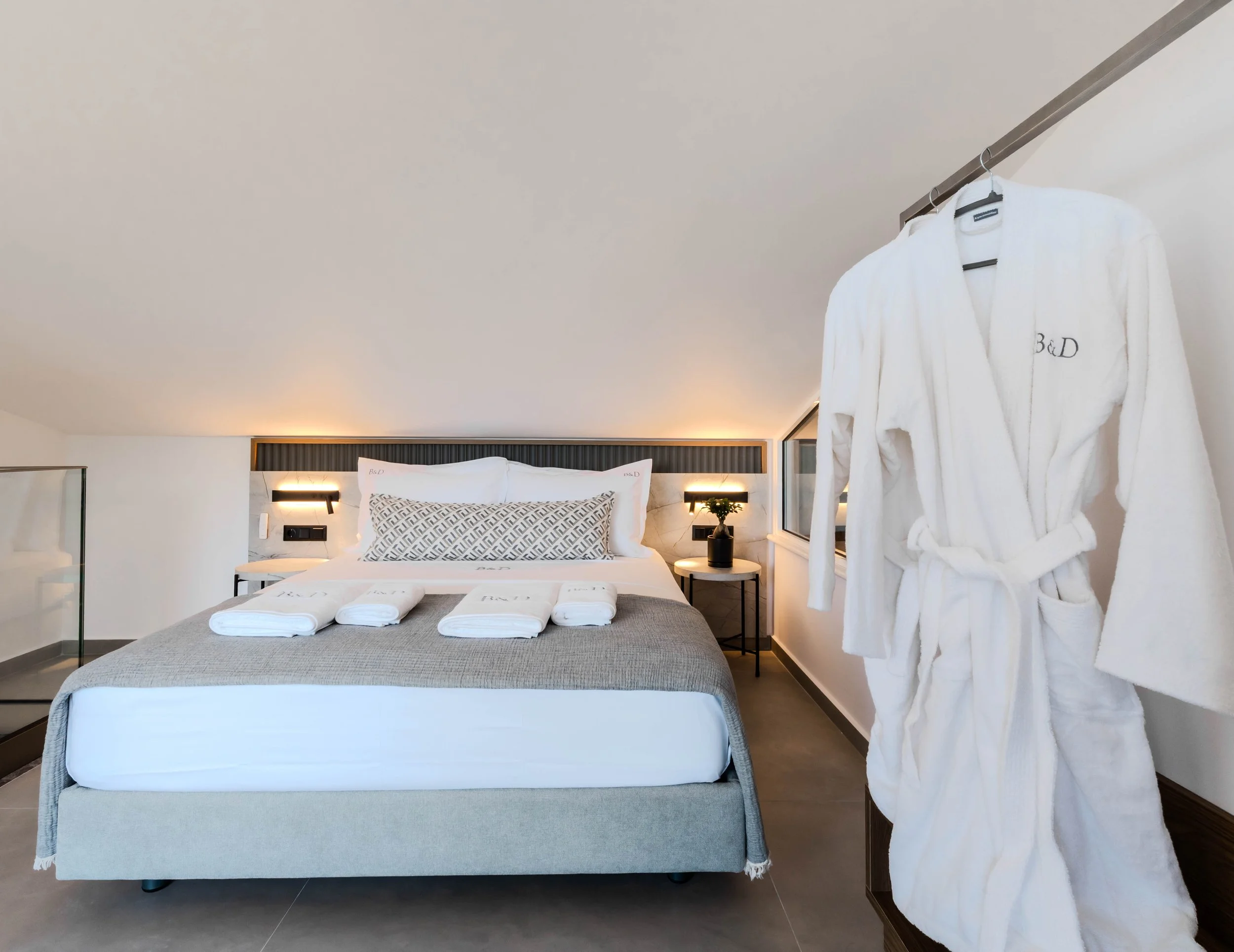 A modern hotel room with a neatly made bed featuring white linens, decorative pillows, and a gray bedspread. Towels are folded on the bed, and a white bathrobe hangs on a rack near the window. The room has minimalistic decor with a side table, a blac
