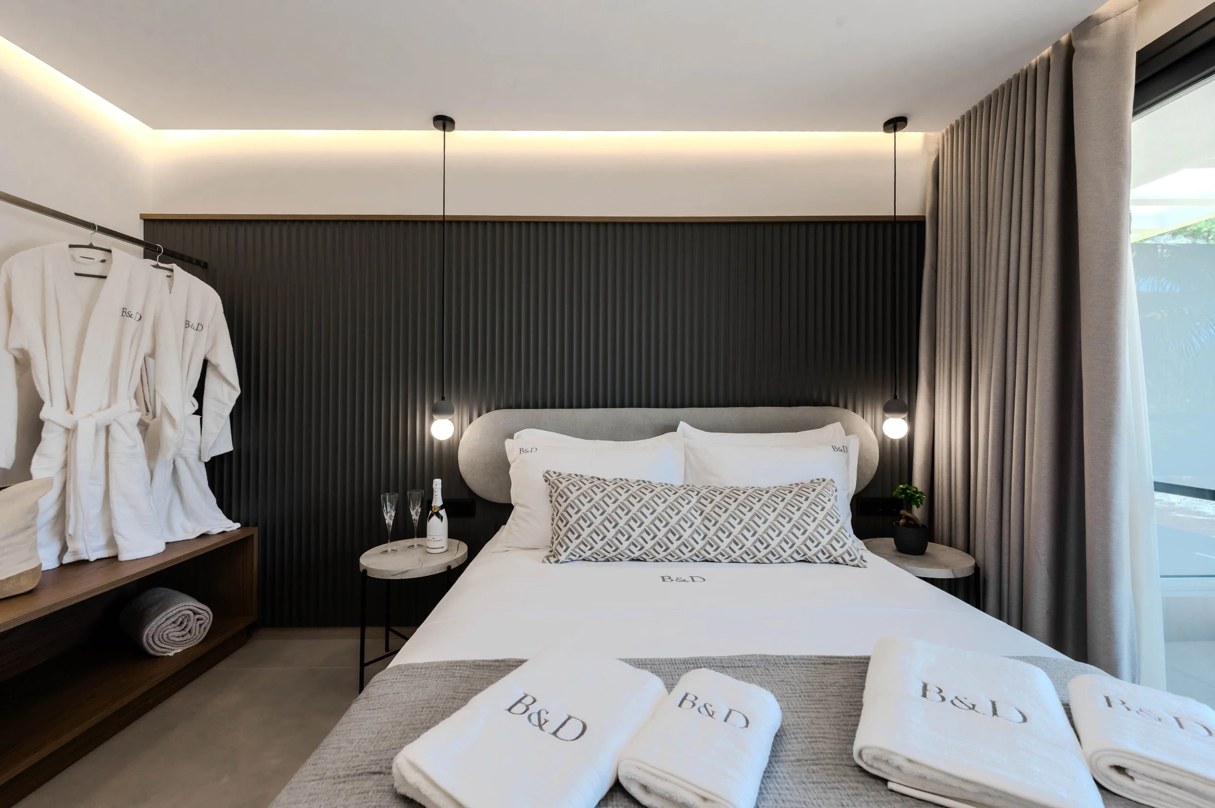 A modern hotel bedroom with a bed, decorative pillows, bedside tables with lamps, bottles of champagne, glasses, and a potted plant, with bathrobes hanging on a rack and white towels on the bed and a window with curtains.