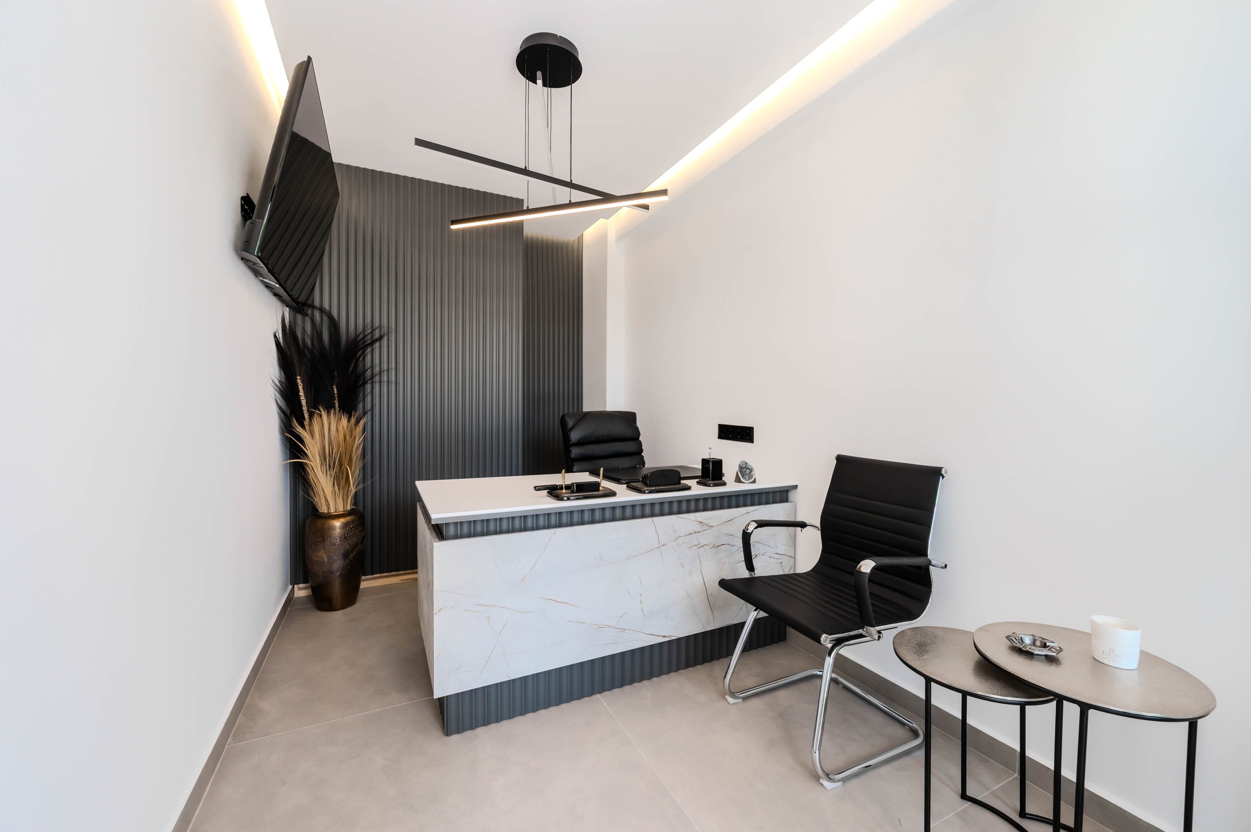 Modern office with white walls, a marble desk, black office chair, and a side table with a white candle and dispensing tray. A black textured accent wall background, a gold and dark vase with dried plants, a wall-mounted TV, and contemporary lighting