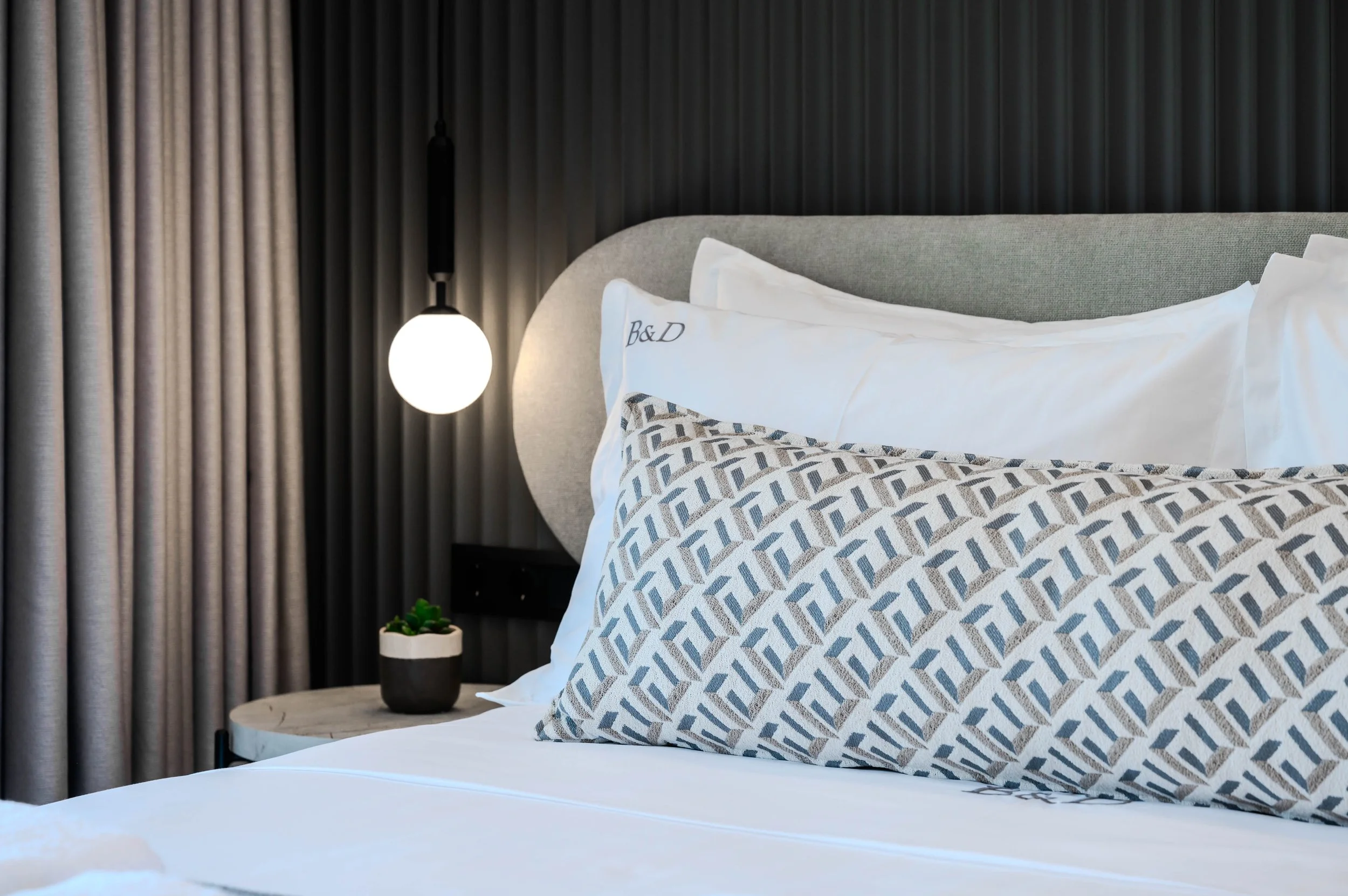 A neatly made bed with white bedding and decorative pillows in a modern hotel room. A small round side table with a plant is beside the bed. A hanging light fixture and dark curtains are in the background.