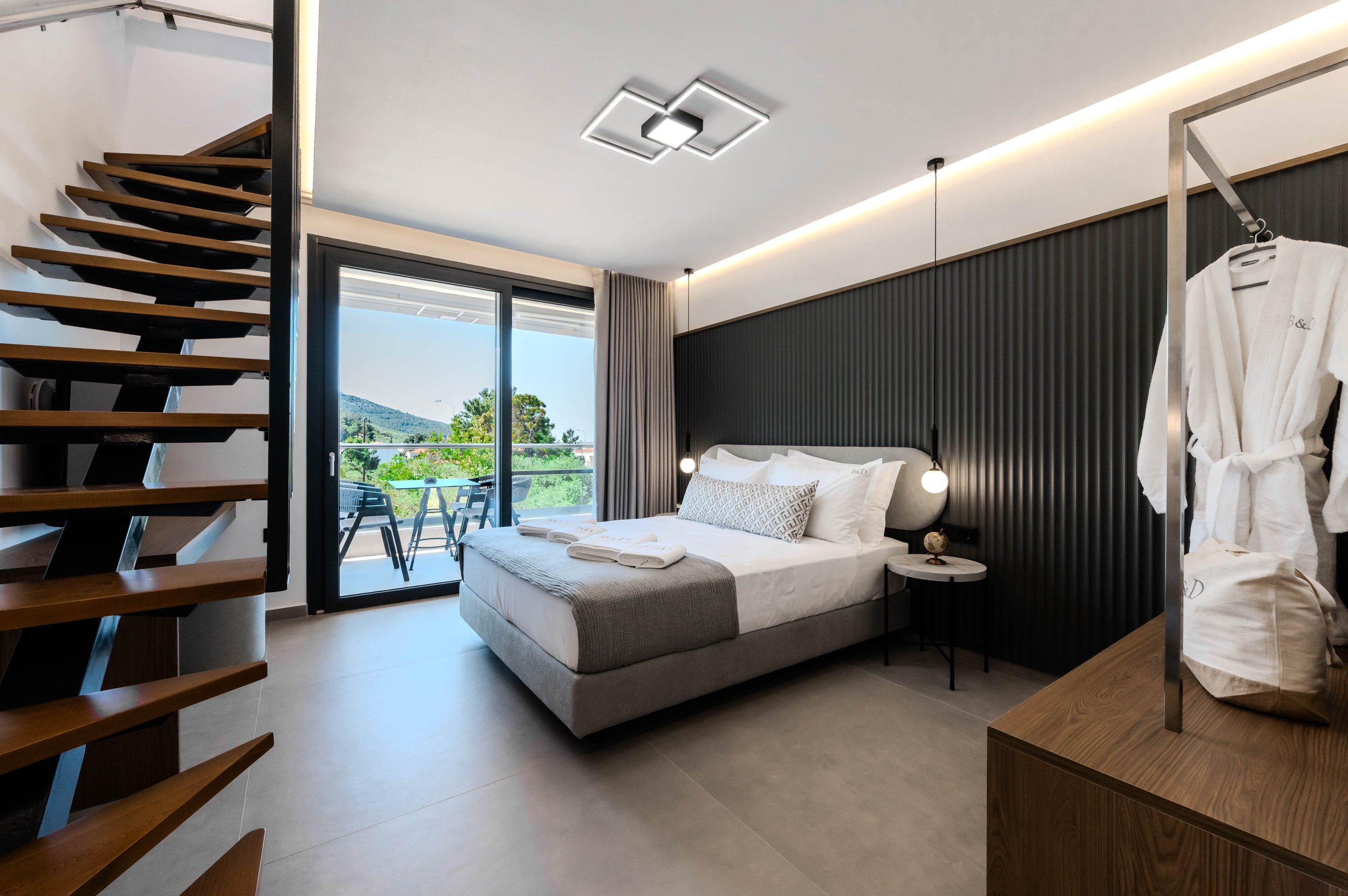 Modern bedroom with a large bed, nightstand, and sliding glass door leading to a balcony with outdoor furniture and scenic view.