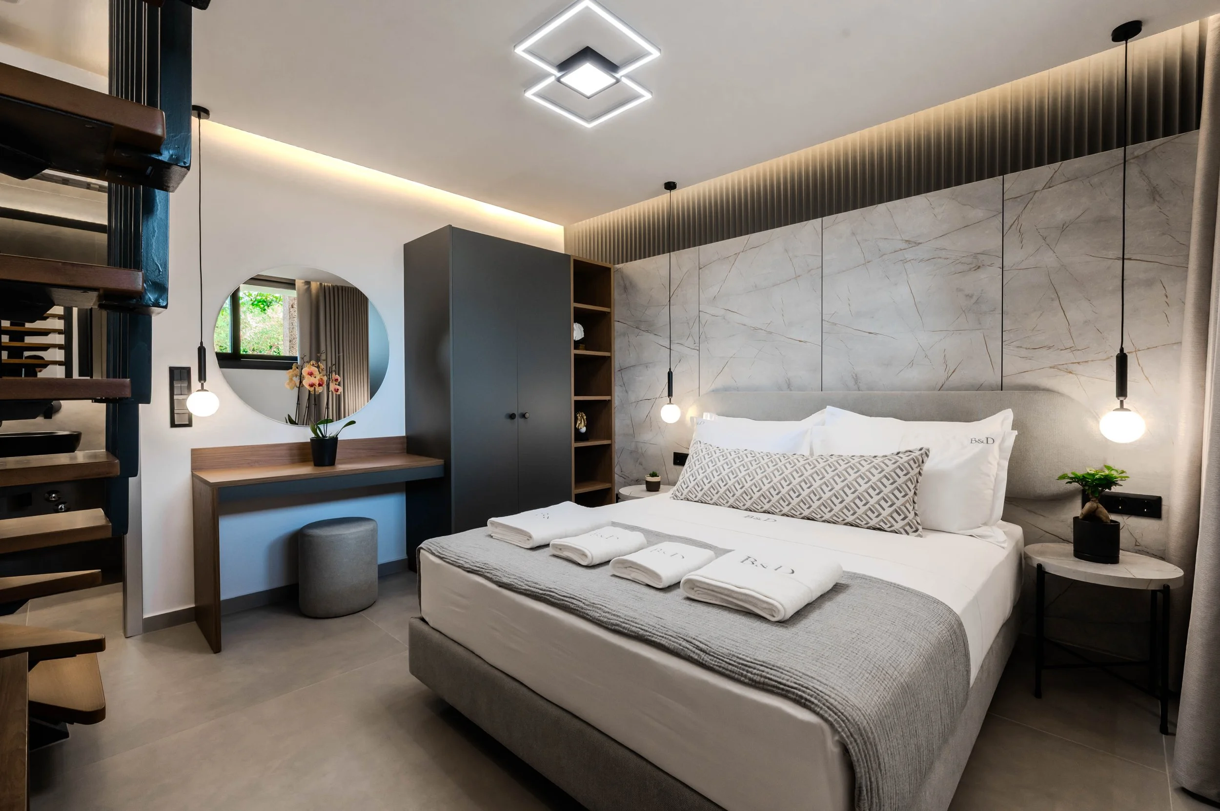 Modern bedroom with a large bed, gray upholstered headboard, white linens, black bedside tables with potted plants, geometric throw pillows, and pendant lights. A vanity with a round mirror, flowers, and a stool is on one side, with a gray wardrobe a
