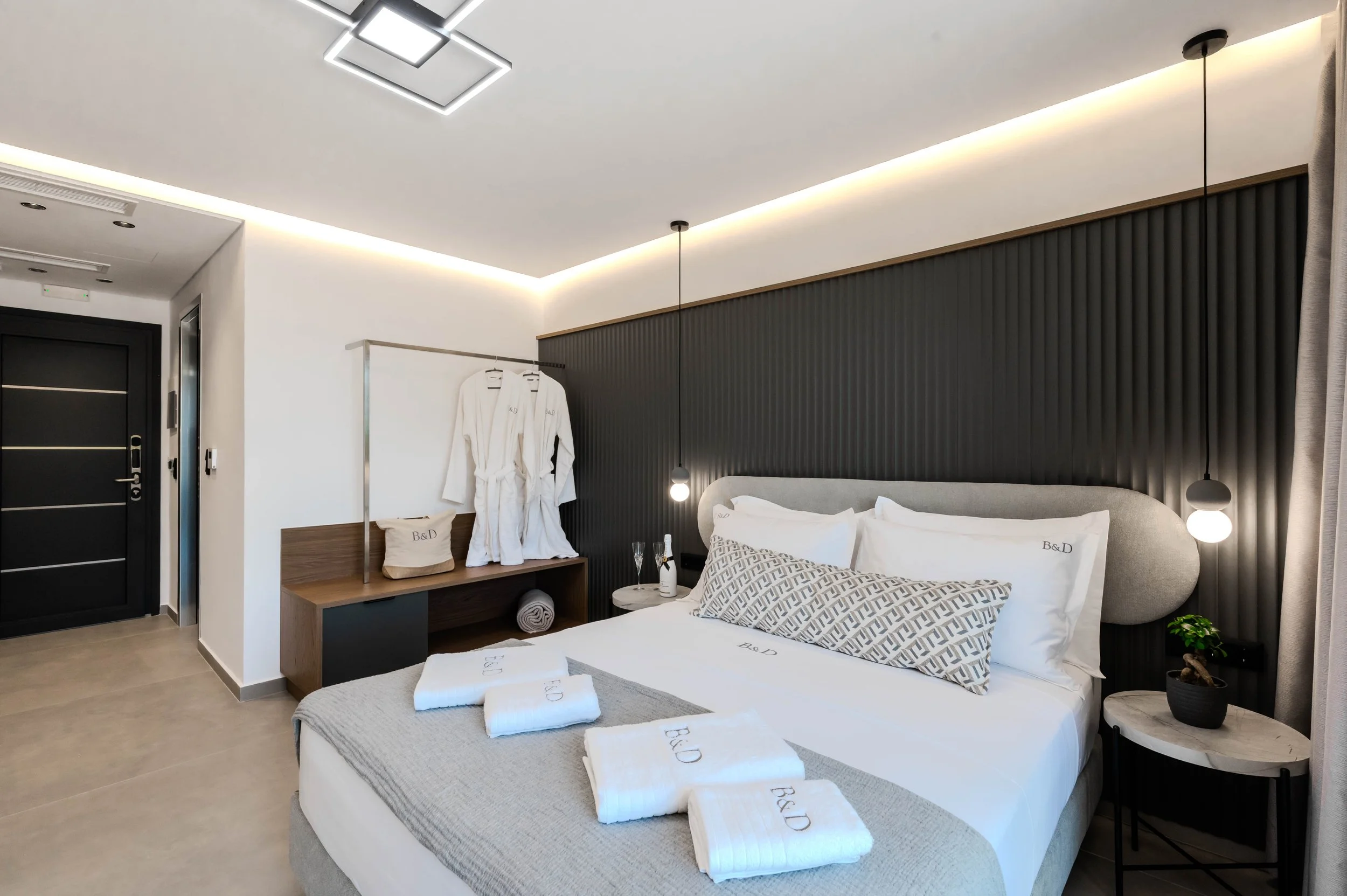 Modern hotel bedroom with a large bed, black textured headboard, white bedding, and decorative pillows, with two pendant lights, a small side table with a potted plant, and a wooden shelf with robes and a bag, near a black door.