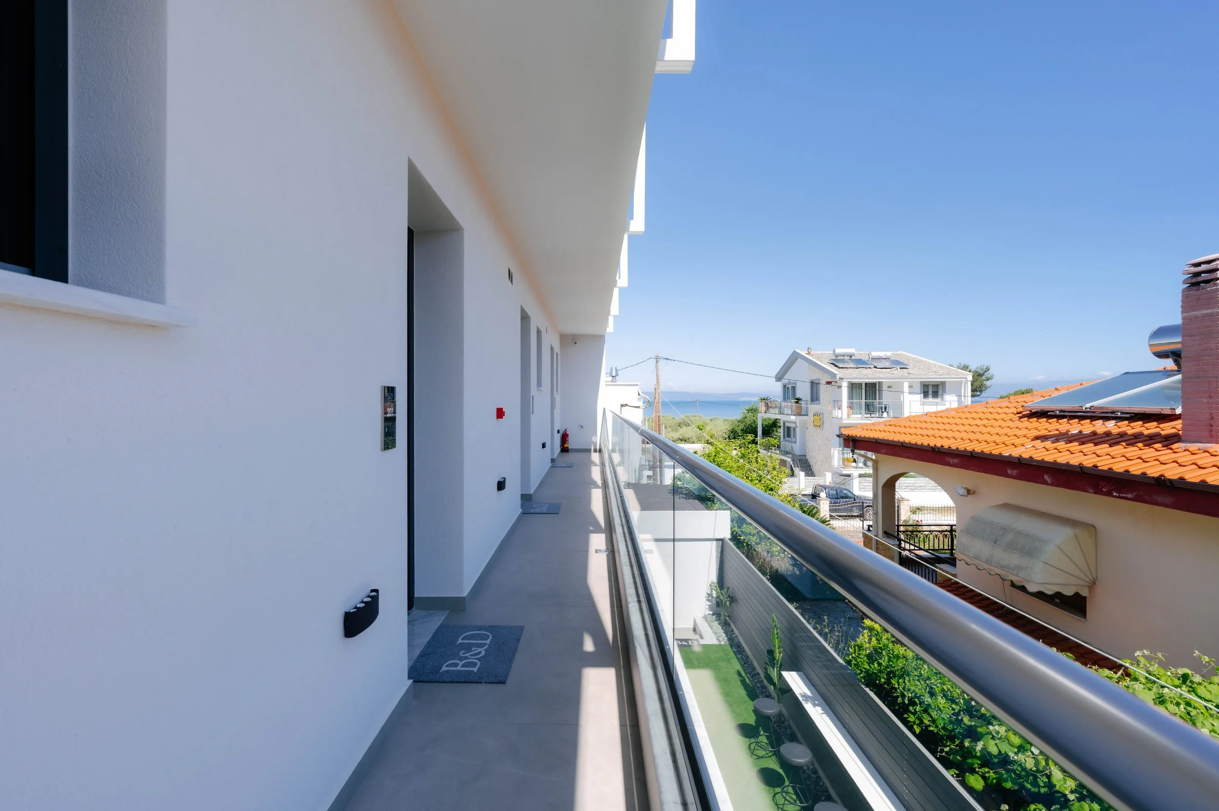 View from a modern apartment balcony looking out over neighboring houses, with a glimpse of the ocean in the distance under a clear blue sky.