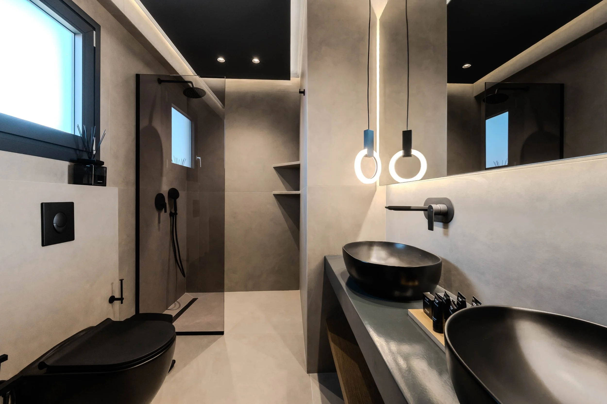 Modern bathroom with black vessel sinks, pendant lighting, black fixtures, a walk-in shower with glass partition, small windows, and minimalist decor.